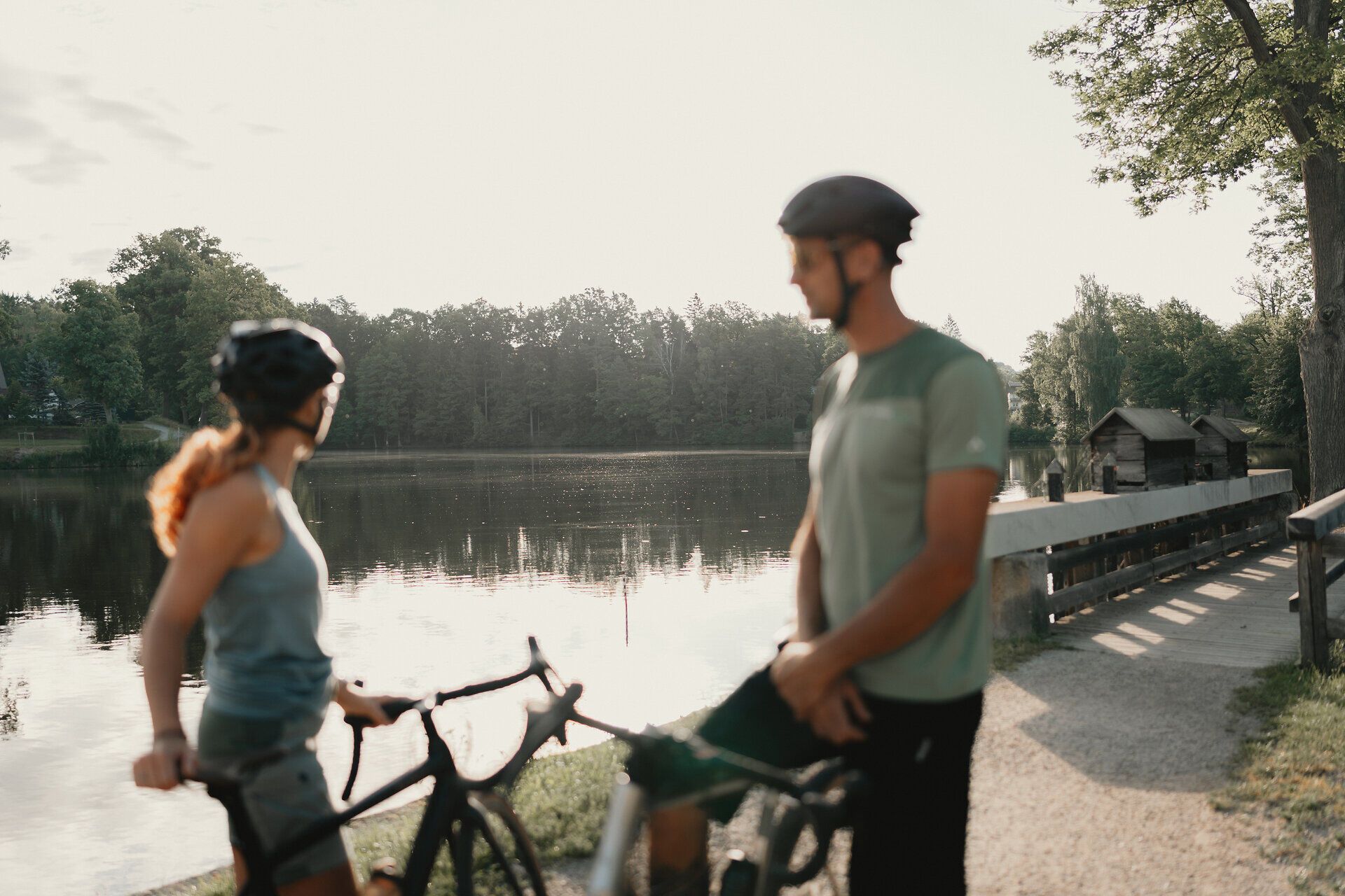 Two cyclists wearing helmets stand with their bikes on the banks of a quiet pond surrounded by forest.