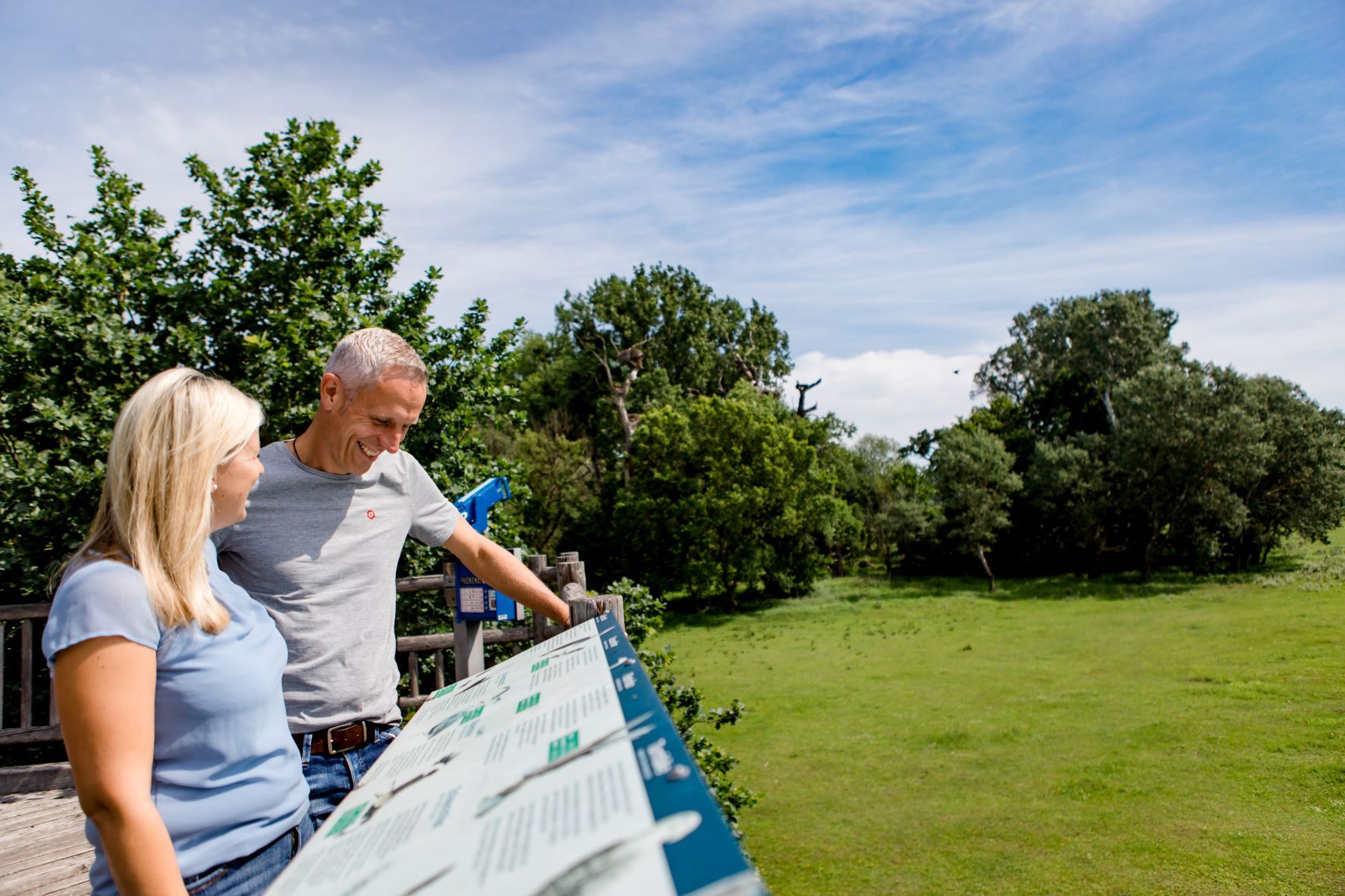 A man and a woman stand on a platform and look at an information board. Trees and a blue sky can be seen in the background.
