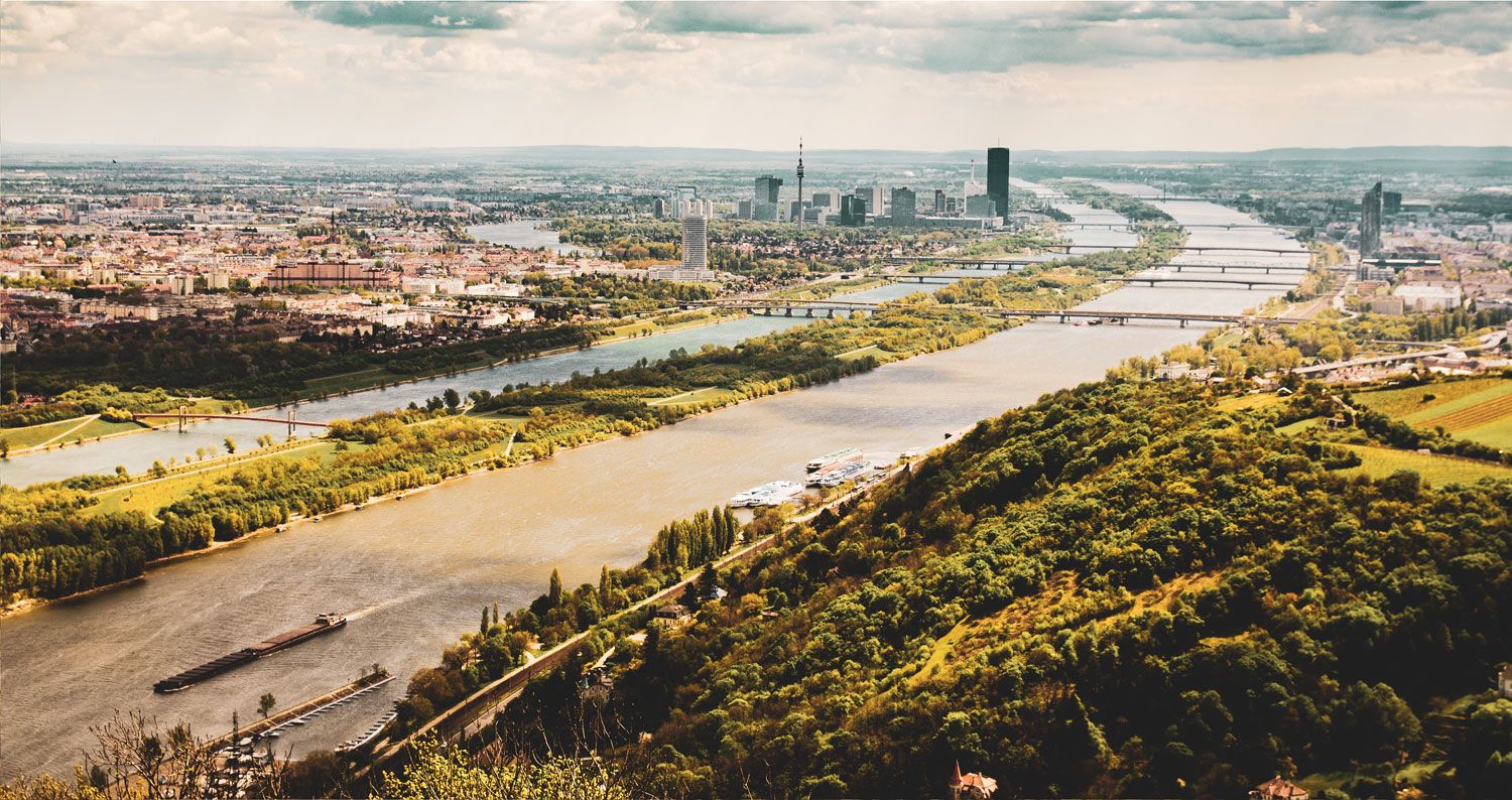 Aerial view of Vienna with the Danube and green hills in the foreground.