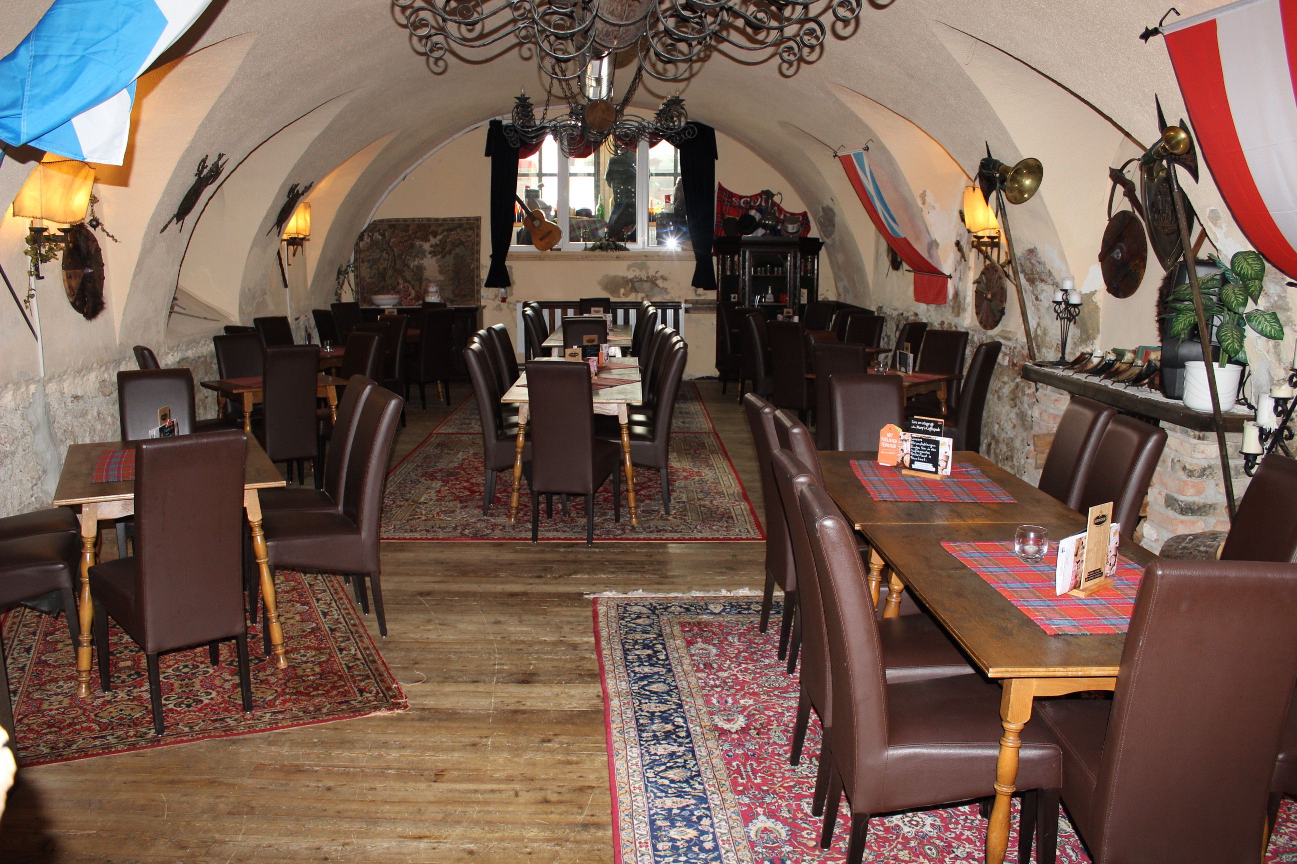 A vaulted hall with tables and chairs, decorated with carpets and flags.