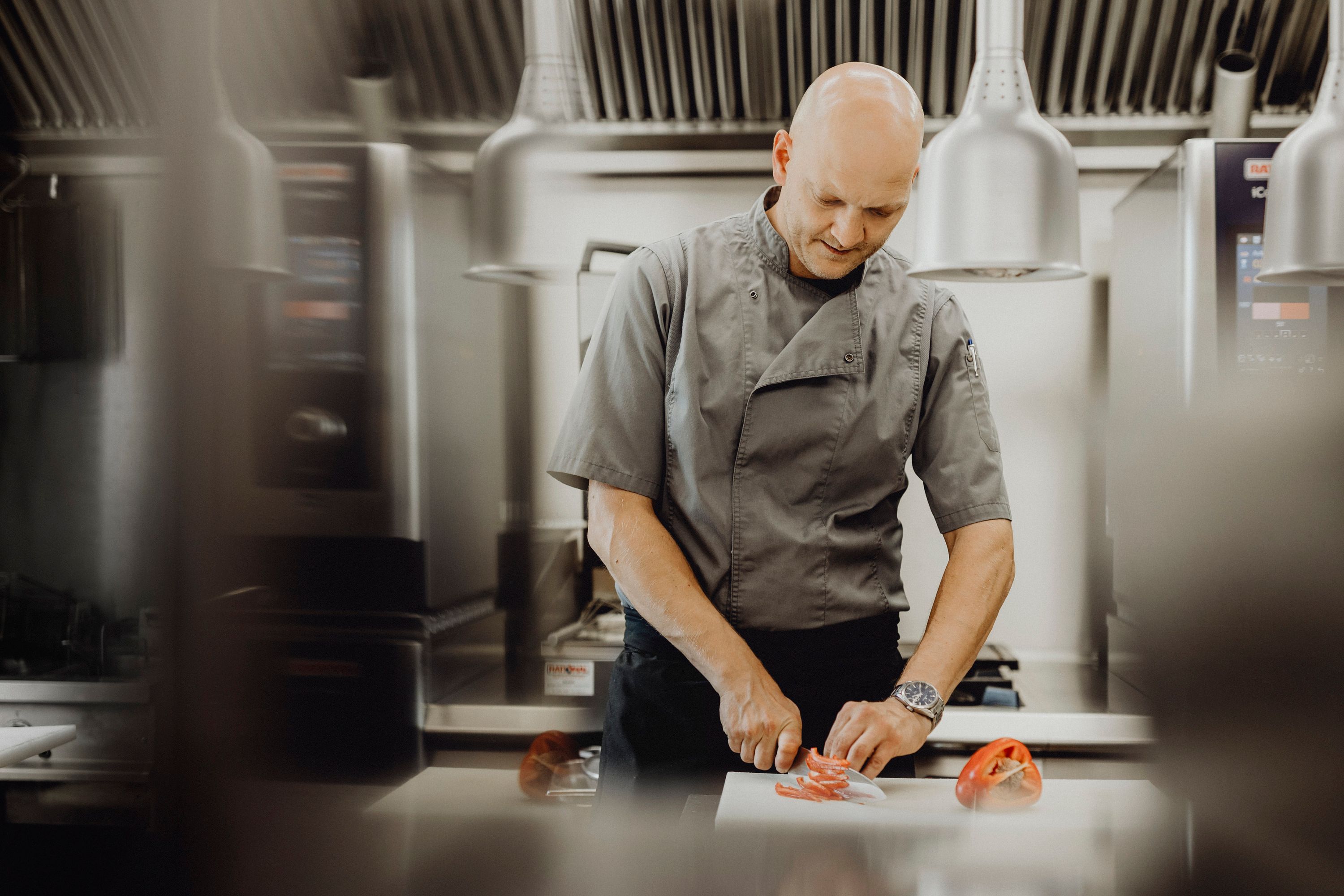 A cook in a gray uniform cuts peppers in a modern kitchen.