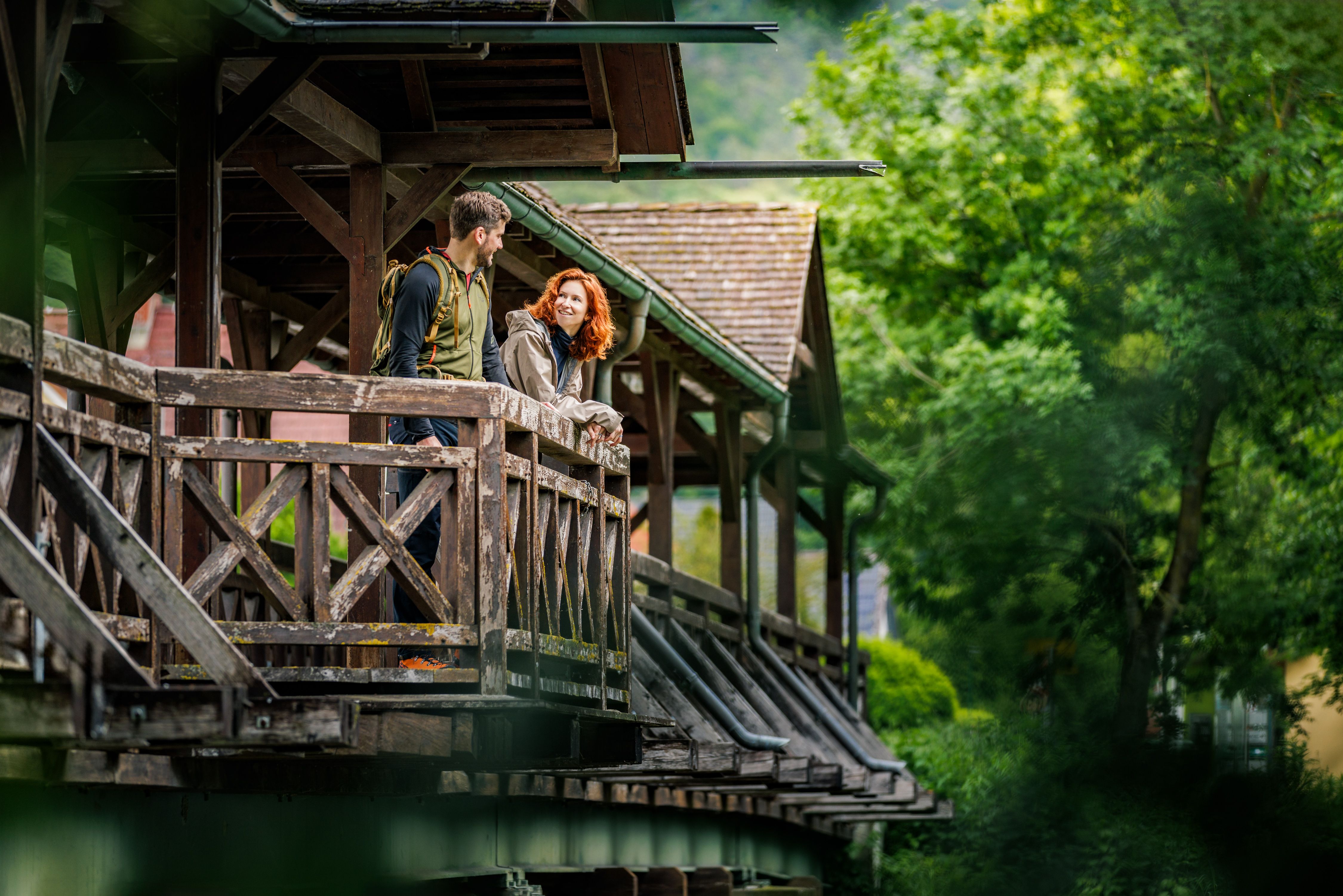 A couple stand on a covered wooden bridge and look at each other, surrounded by green nature.