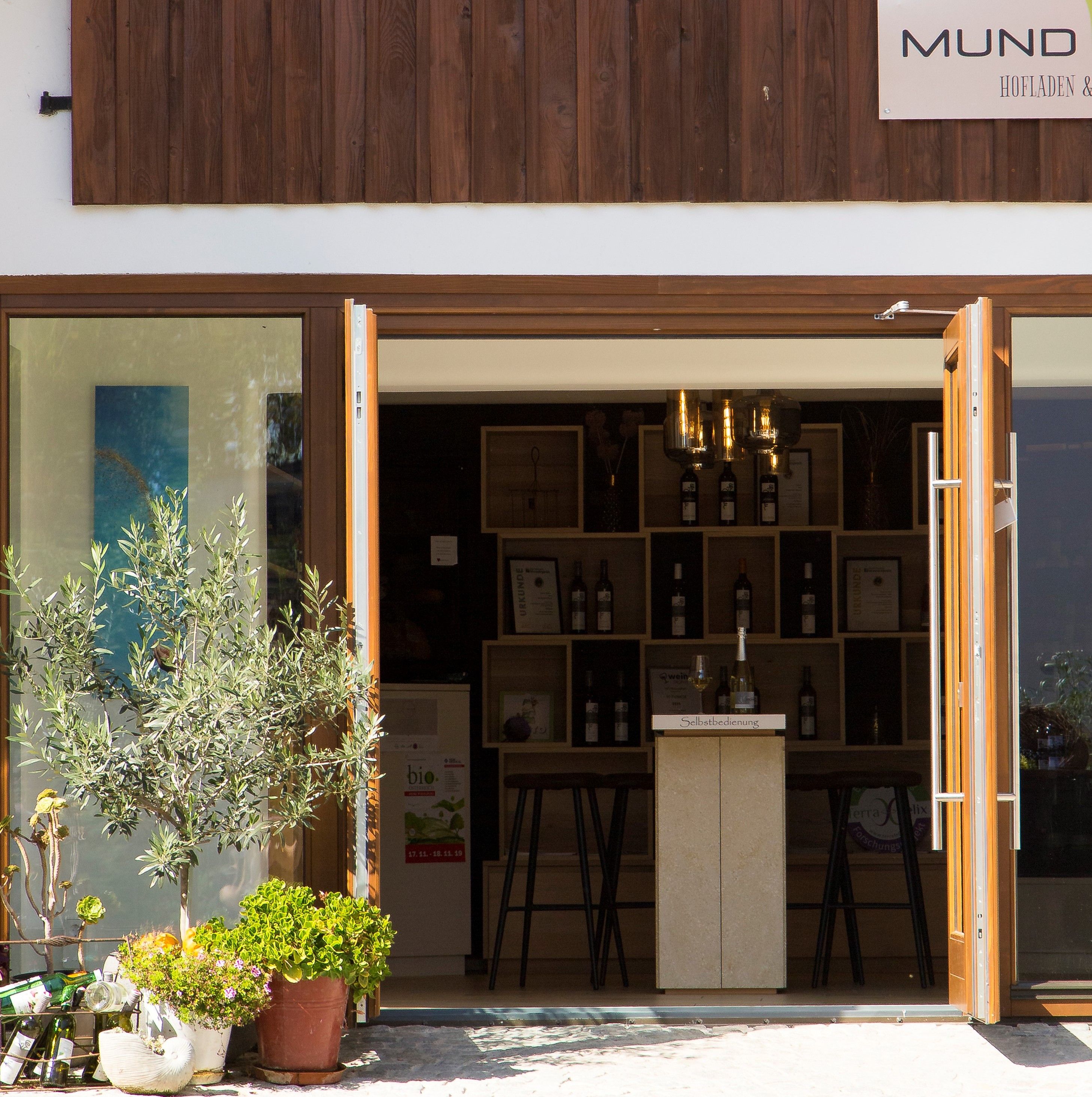 Entrance to a farm store with an open wooden gate, plants and bottles of wine.
