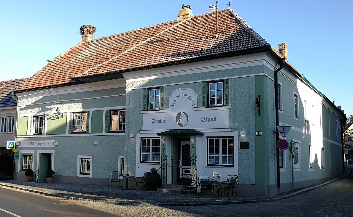 A traditional inn with a green façade and the name 'Zum weißen Rössl'. There are tables and chairs in front of the building.