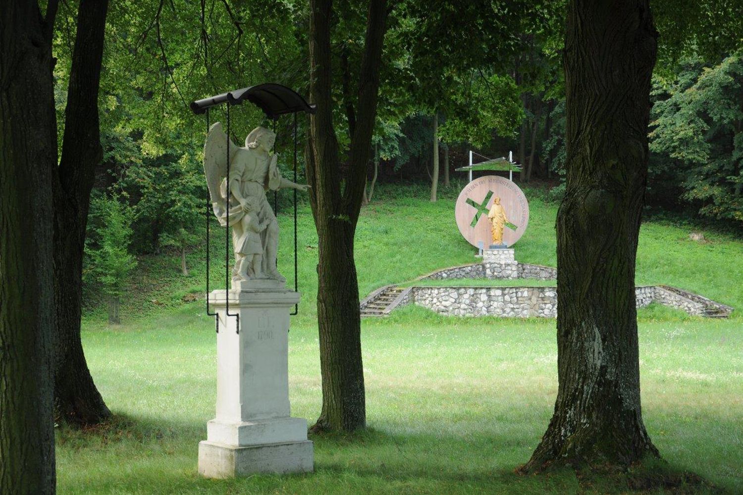 Statue of an angel with child in the foreground, in the background another statue on a pedestal in a wooded park.