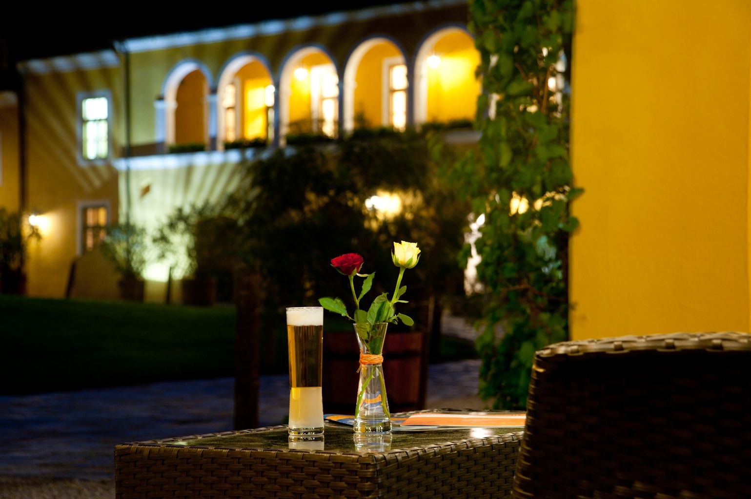 A table with a glass of beer and two roses in a vase, in the background an illuminated building with arches.