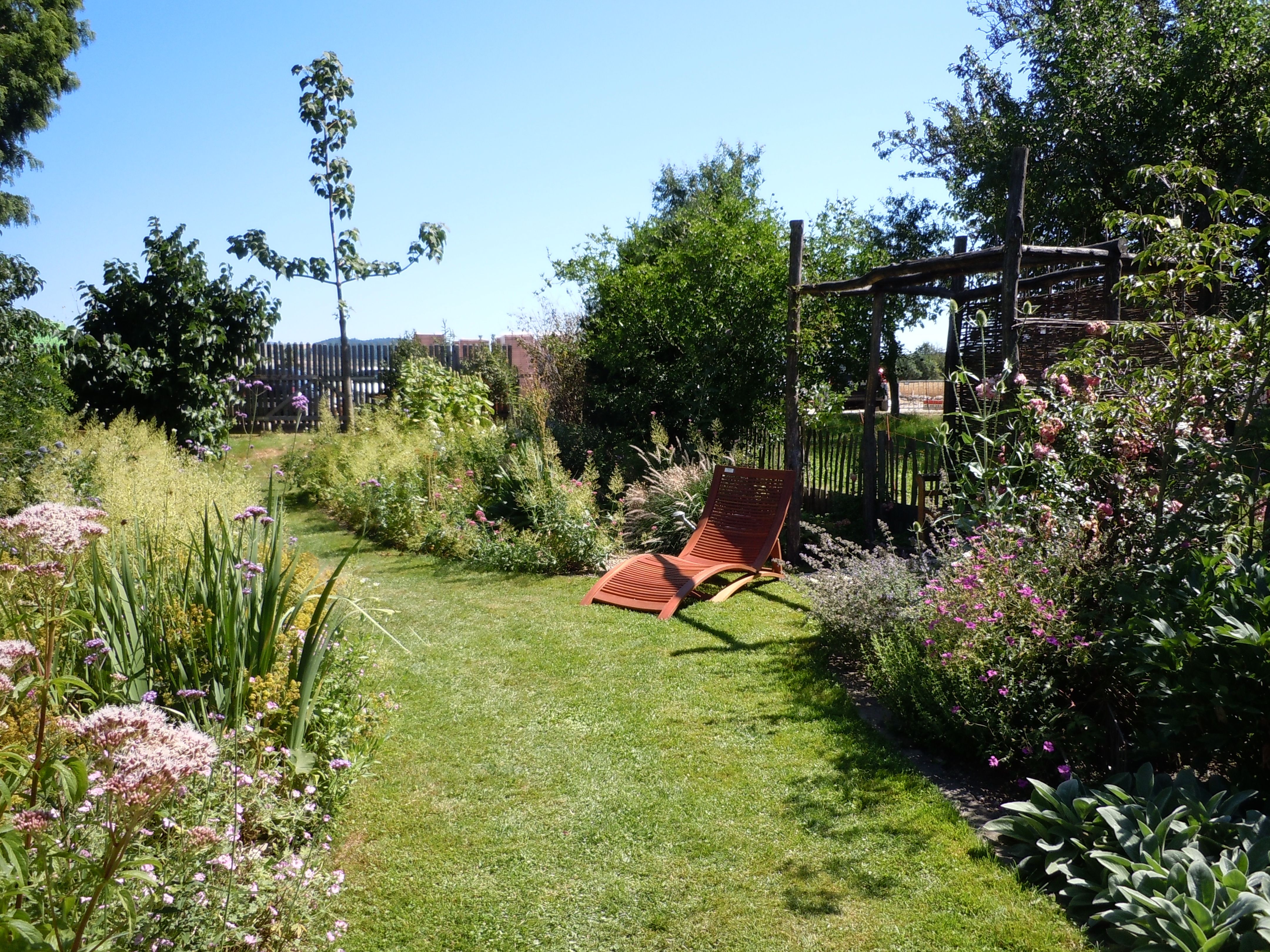 A well-tended garden with sun loungers, surrounded by flowering plants and trees under a blue sky.