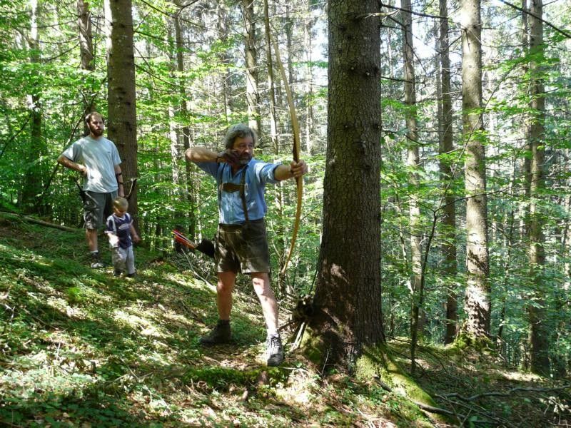 Person shoots with a bow in the forest, two other people in the background.