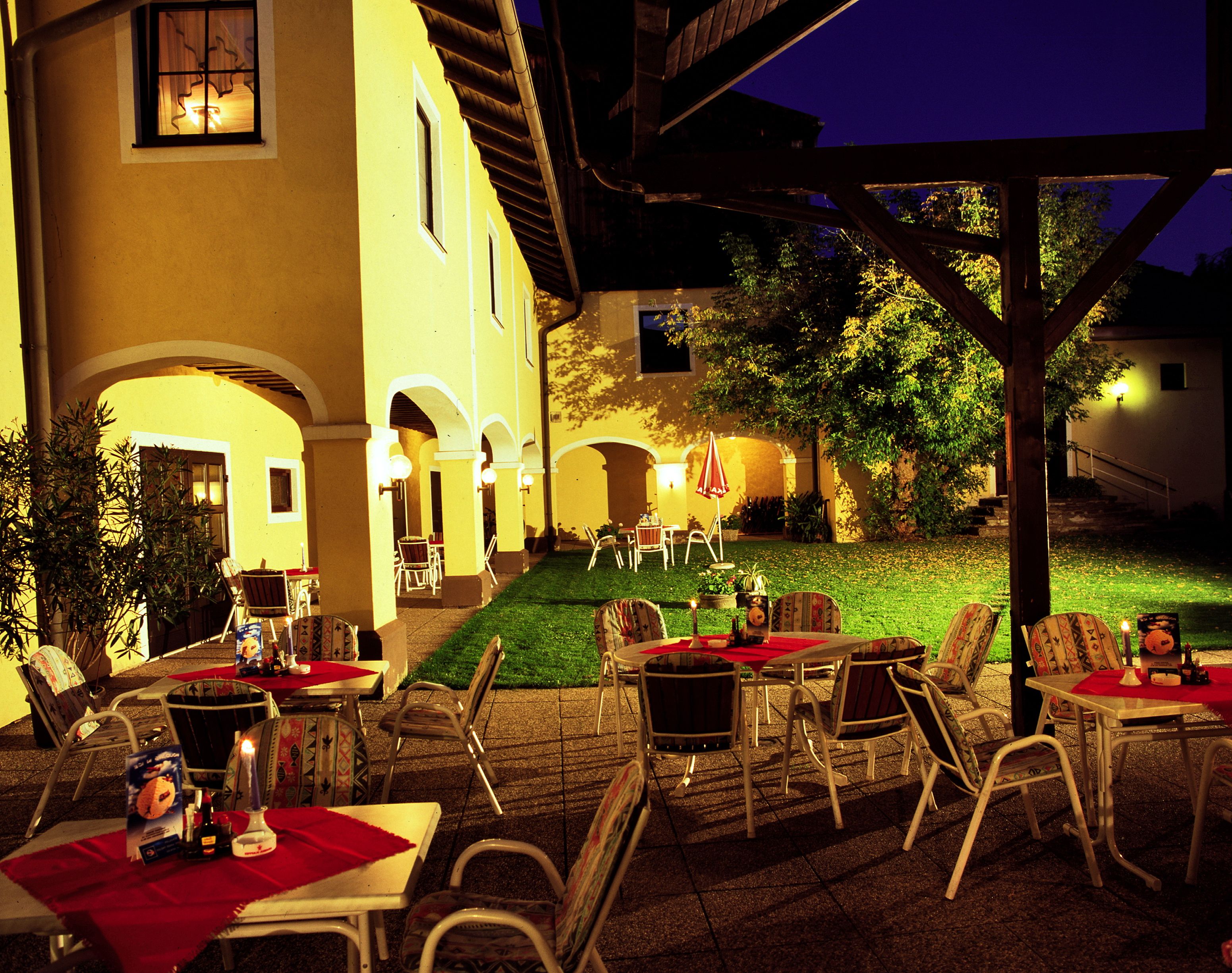 Illuminated outdoor area of an inn with yellow walls, tables and chairs on a terrace at night.
