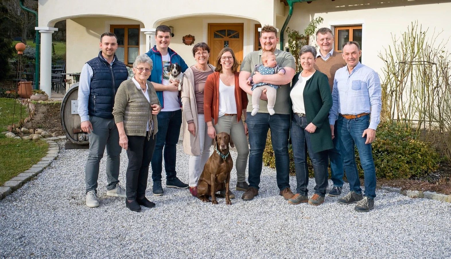 A family stands in front of a house, four generations together with a dog and a baby.