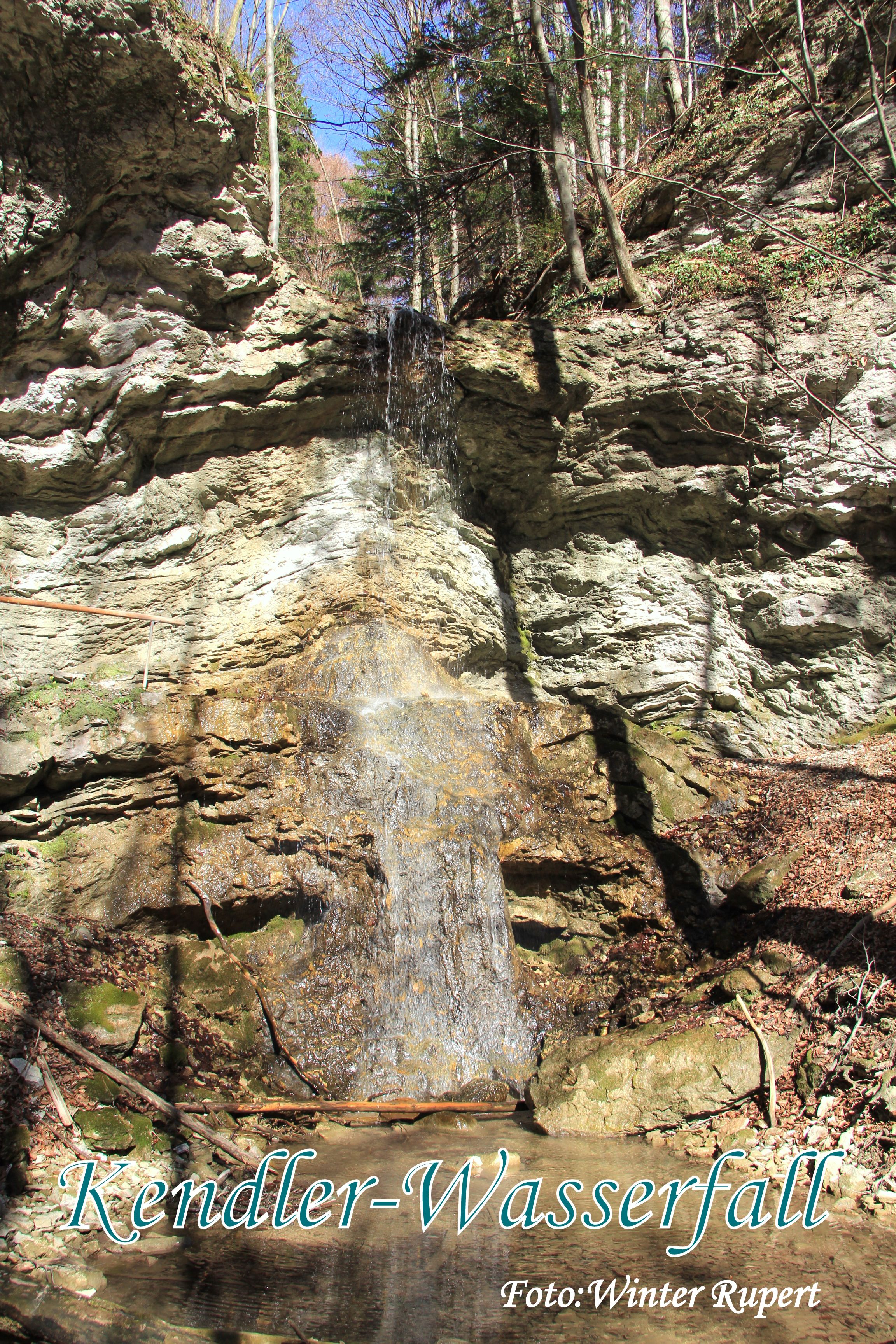 A small waterfall flows over a rock face, surrounded by trees and rocks.