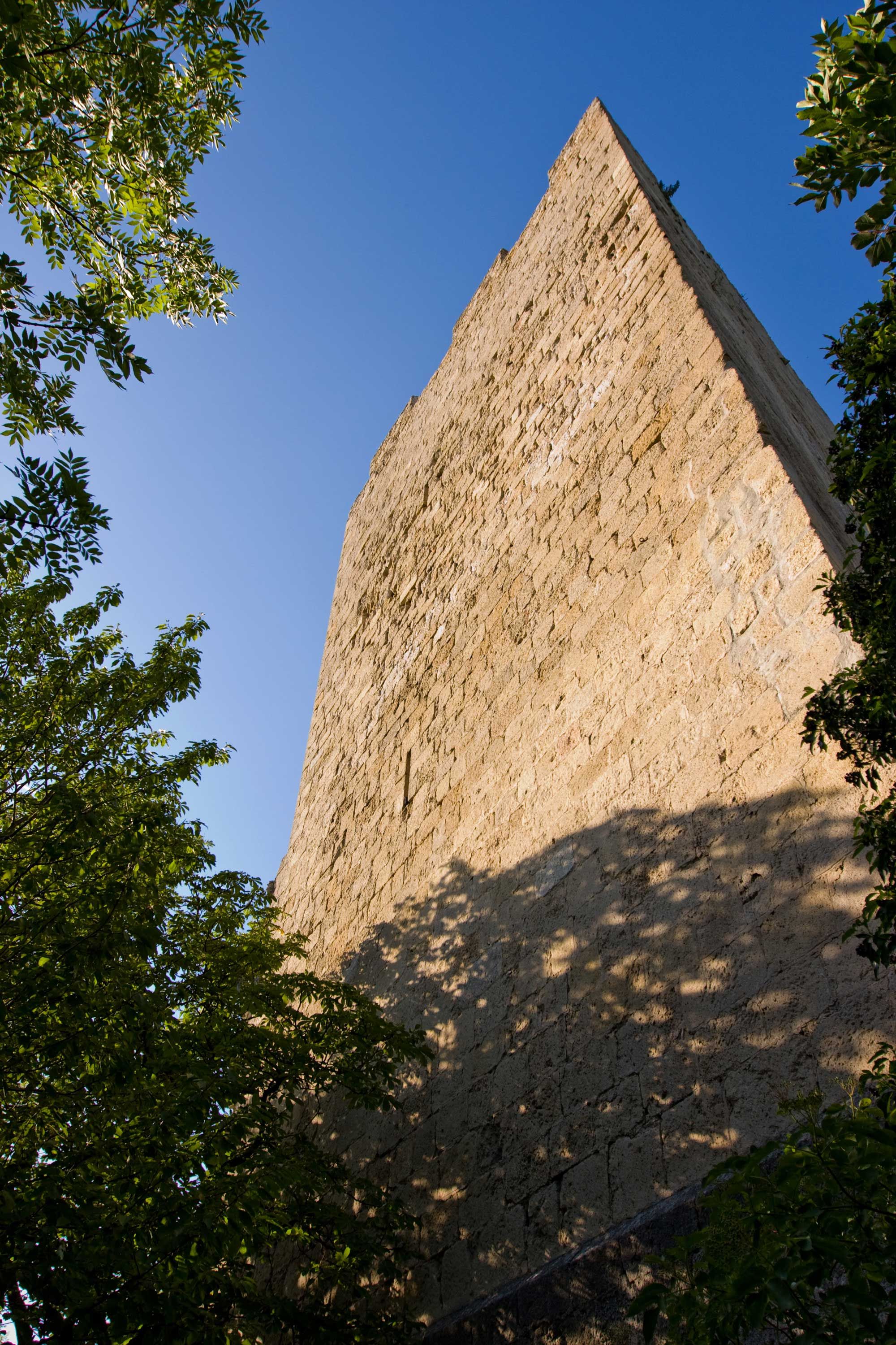 Close-up of a tall, old stone castle tower, surrounded by trees, under a blue sky.