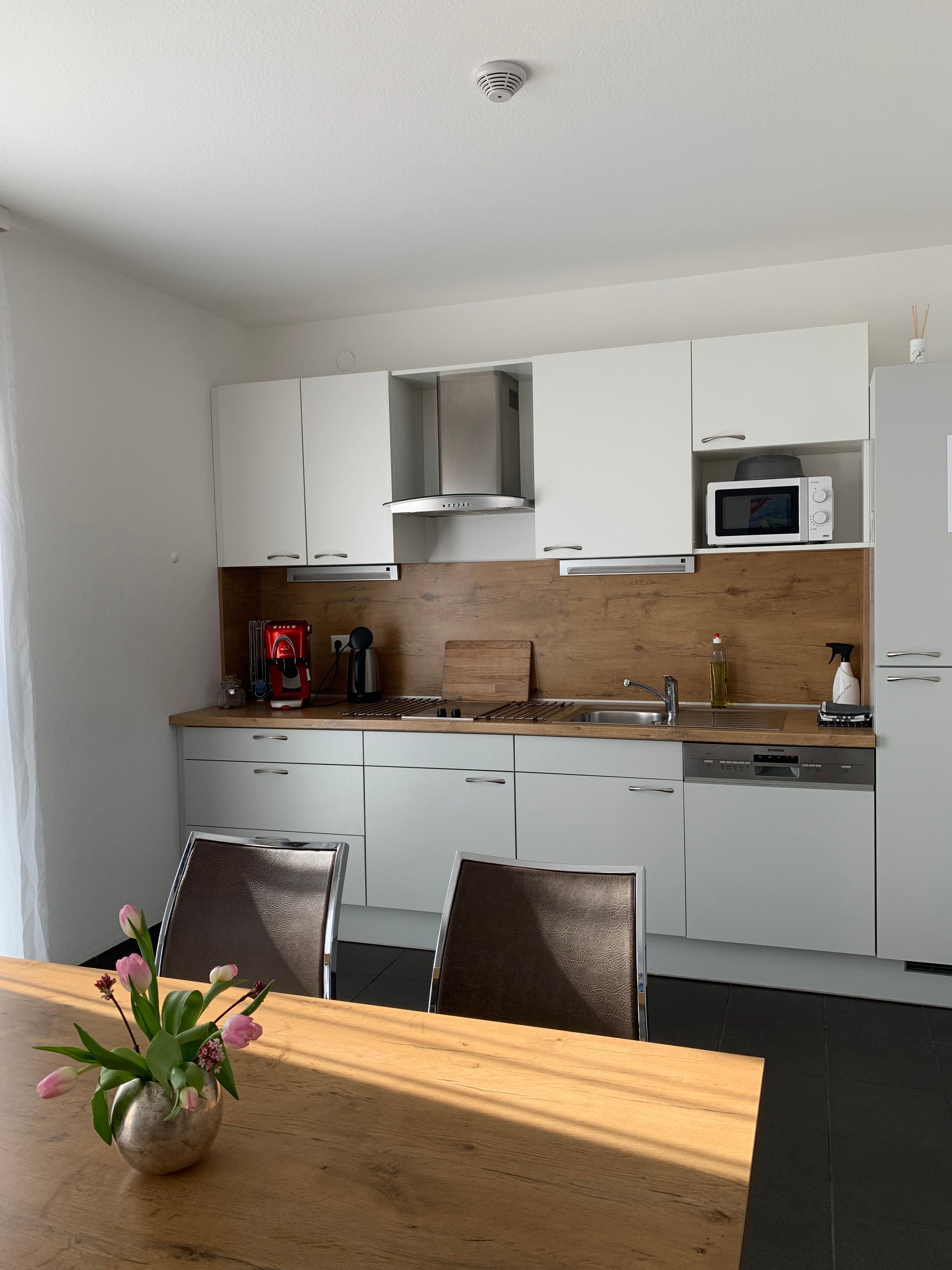 Modern kitchen unit with white cupboards, wooden worktop, coffee machine and microwave. A table with a bouquet of flowers in the foreground.
