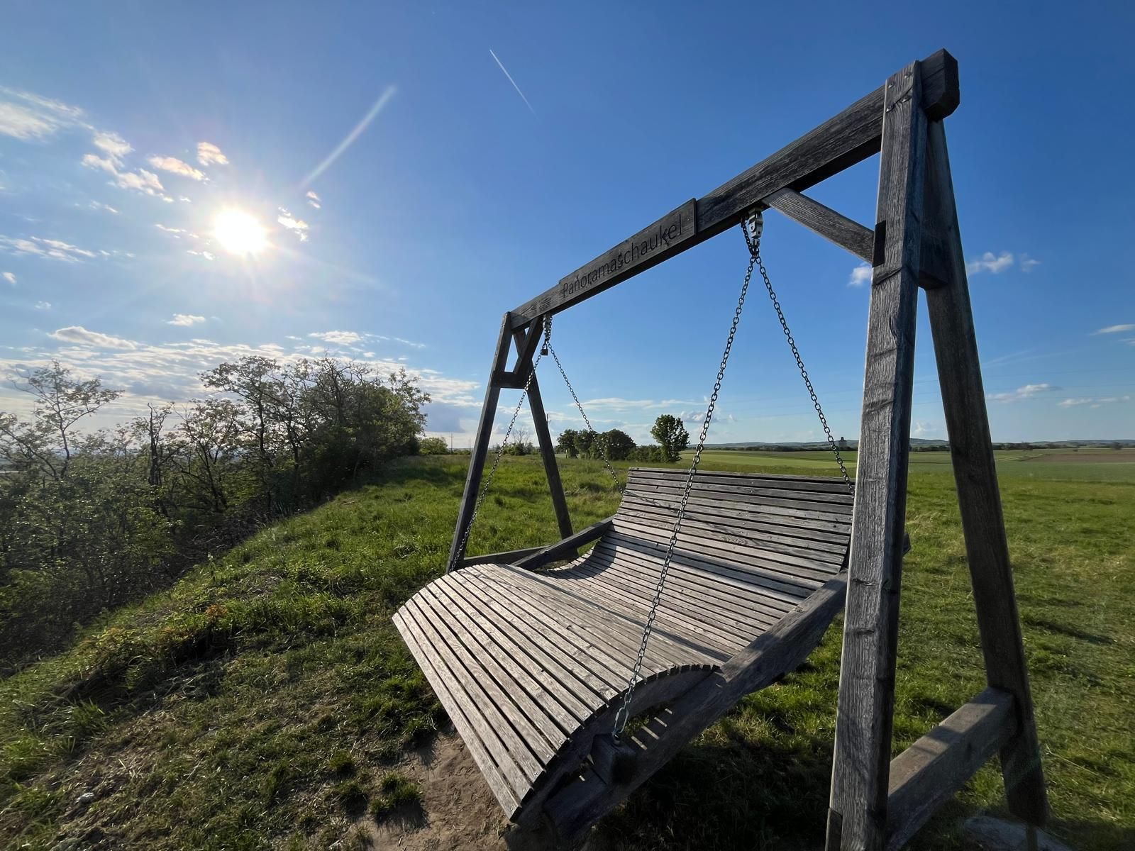 Large wooden panoramic swing on a green meadow under a bright blue sky. The low sun illuminates the landscape and casts long shadows, while individual clouds and a vapor trail cross the sky. The scene appears calm and inviting with a wide view over fields and nature.