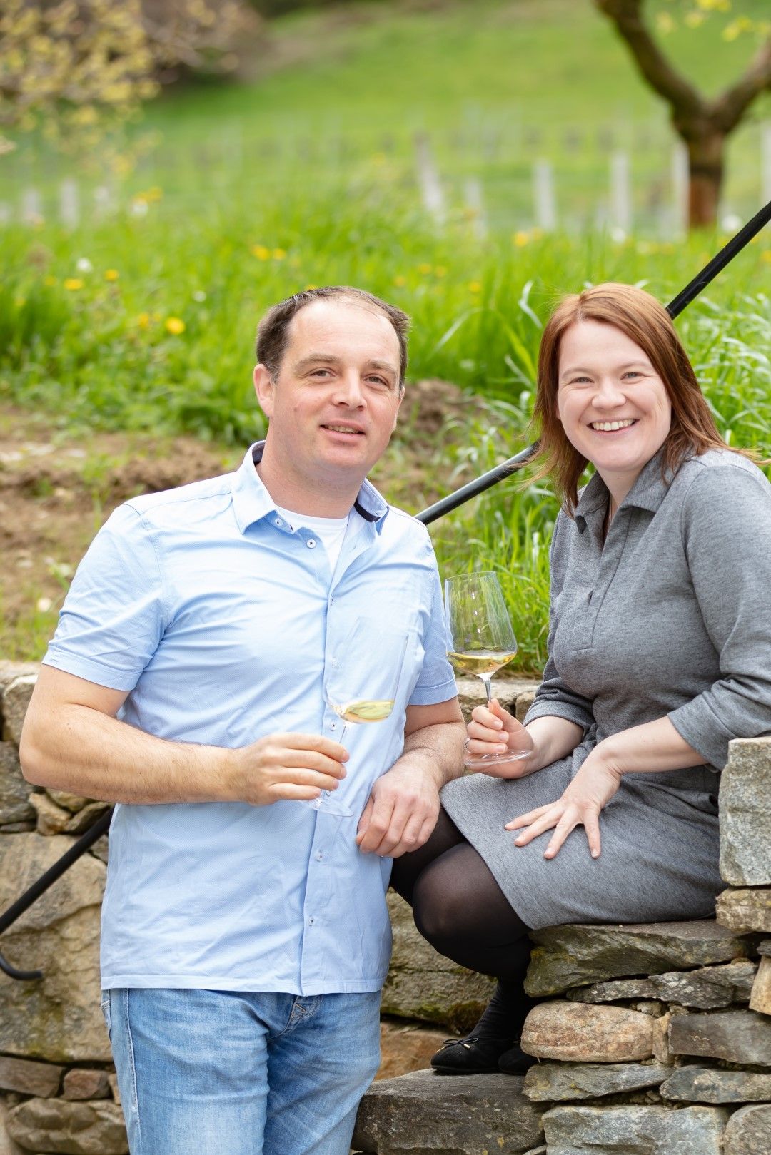 A man and a woman are sitting on a stone wall outside, holding glasses of wine.