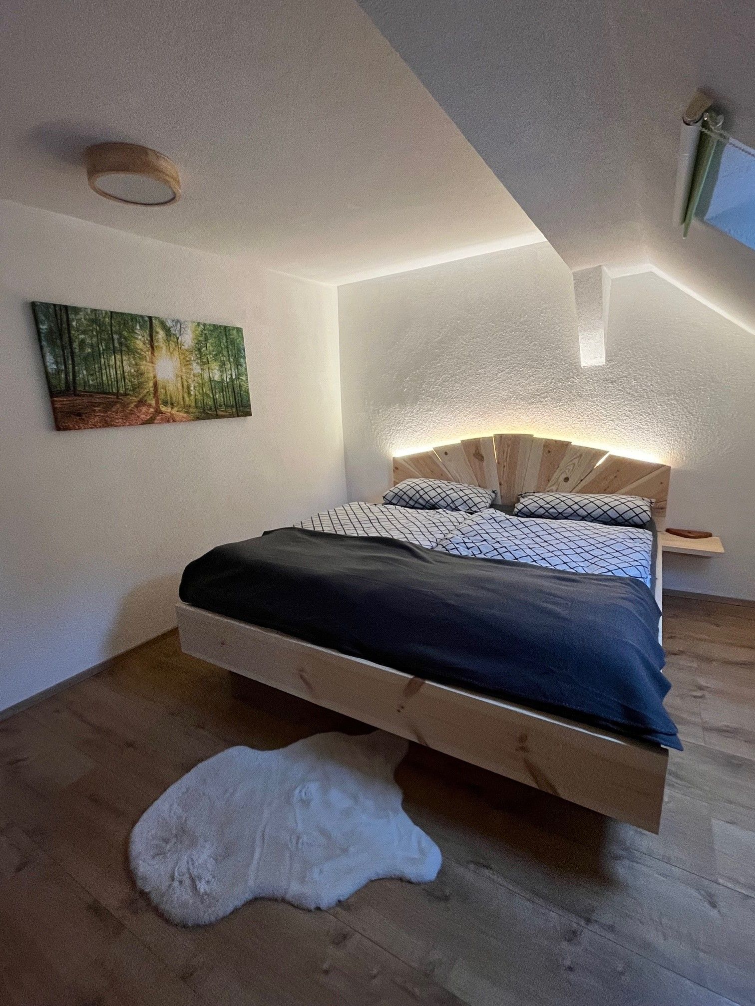 A cozy bedroom with a wooden bed, a picture of a forest on the wall and a white carpet on the wooden floor.