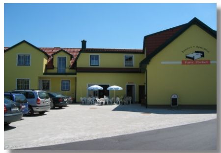 Yellow building with the sign 'Pension Ziekel', cars and tables with parasols in front of it.