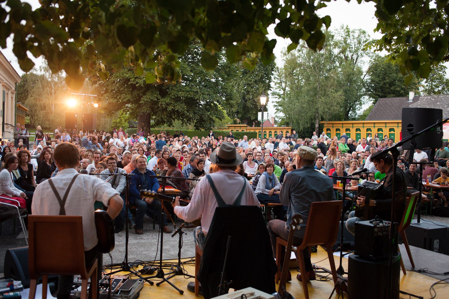 Open-air concert with musicians on a stage and a large outdoor audience.