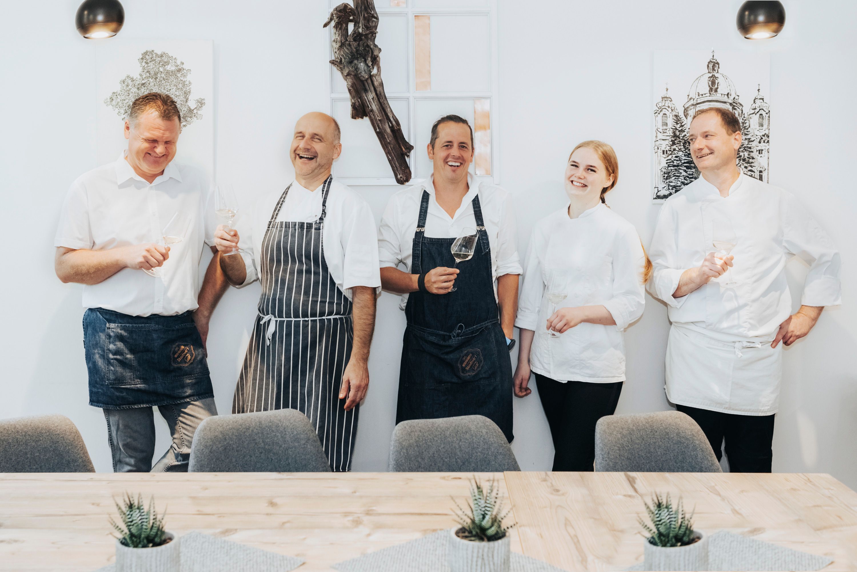 Five people dressed as chefs are laughing and holding glasses of wine.