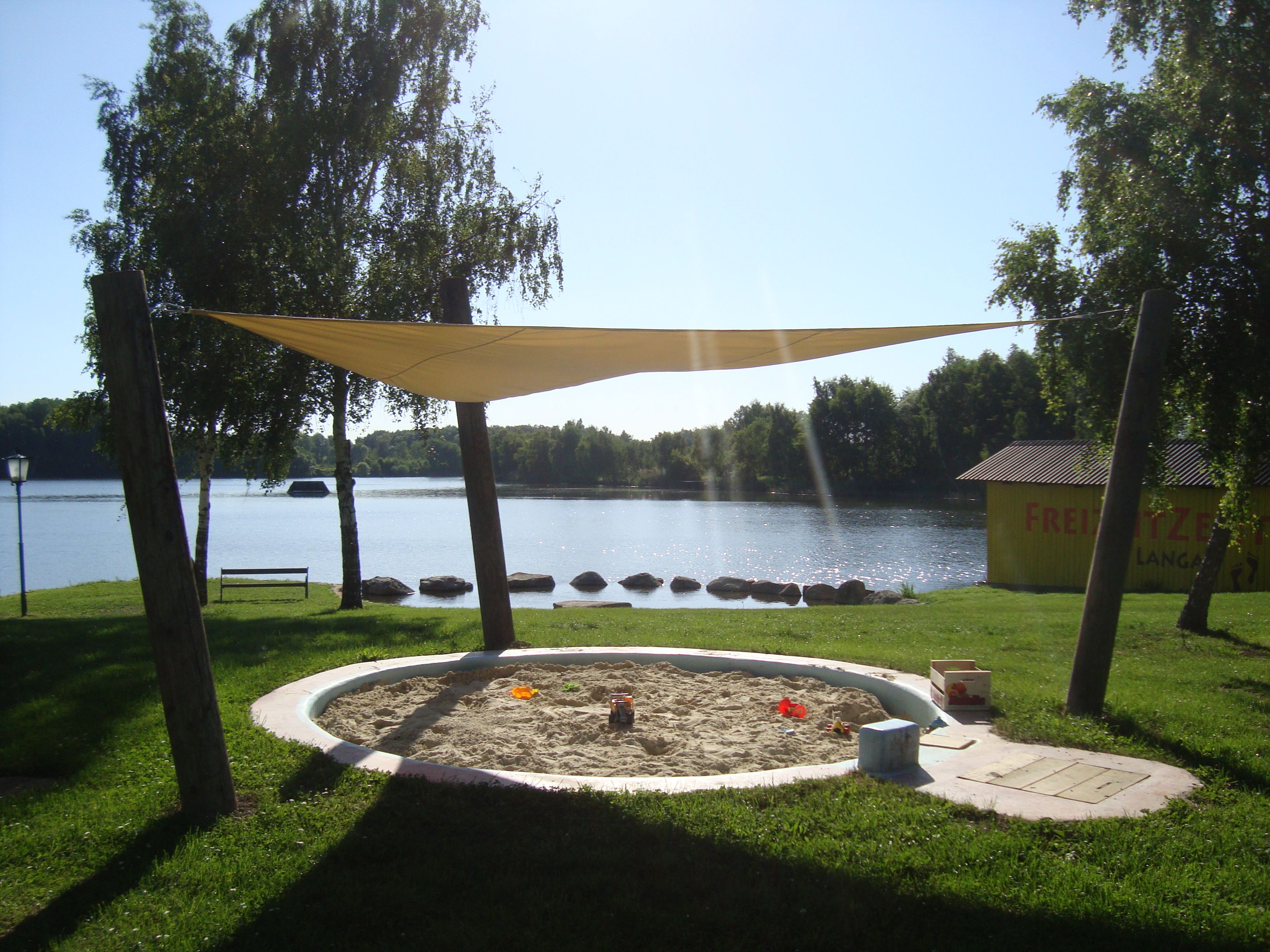 A sandpit with a sun sail on the shore of a lake, surrounded by trees and a meadow. In the background a yellow building with the inscription 'Freizeitzentrum Langau'.