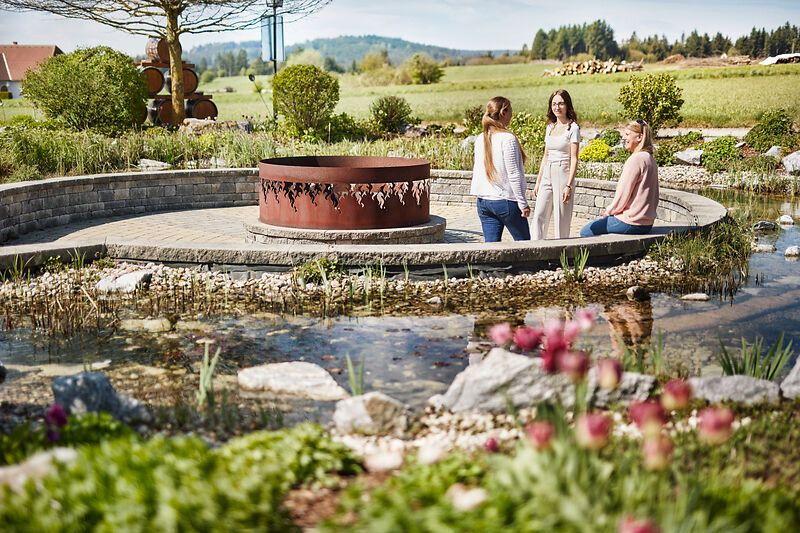 Three women sit in a garden with a small pond and a metal sculpture in the middle.