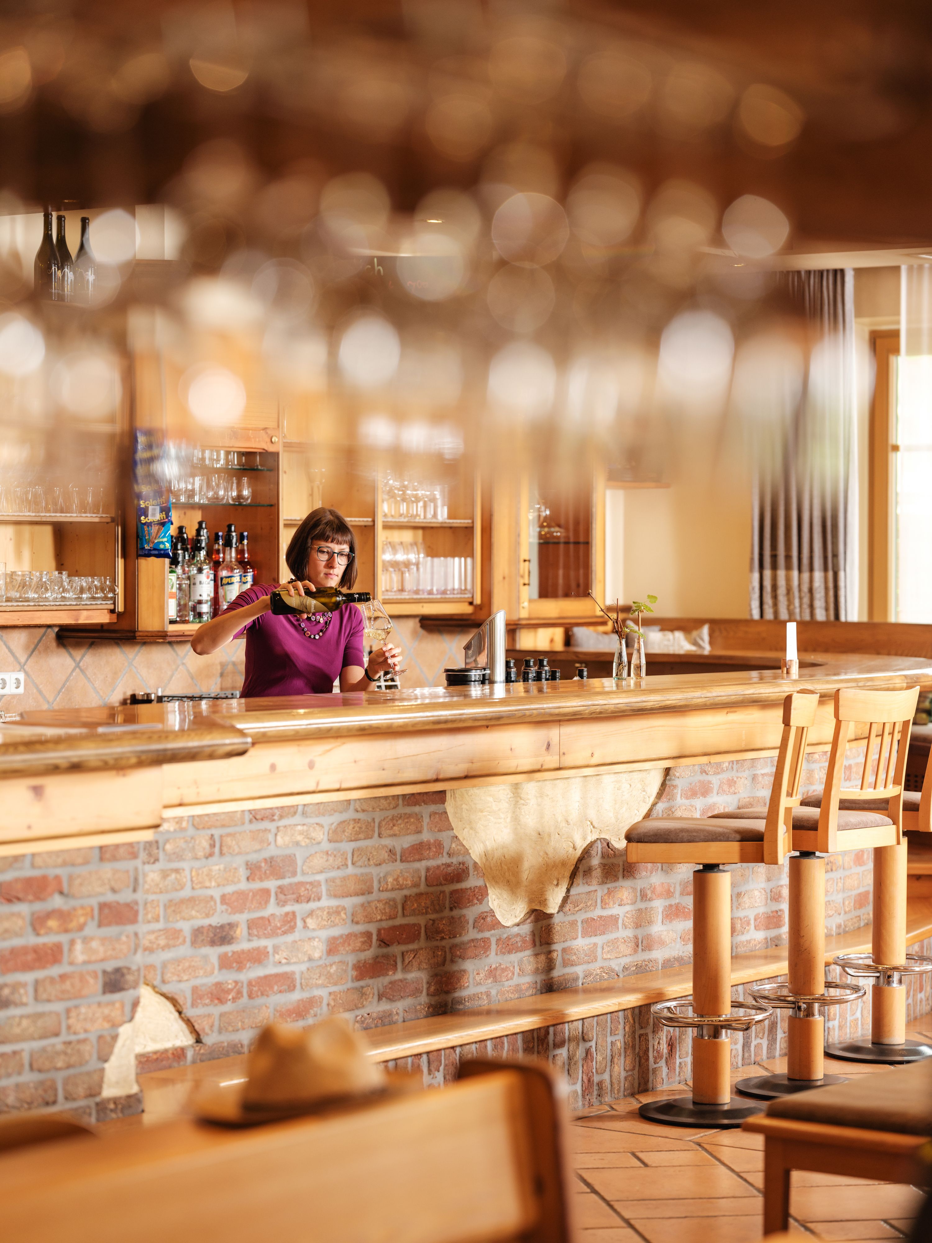 A woman stands behind a bar in a rustic restaurant and pours wine.