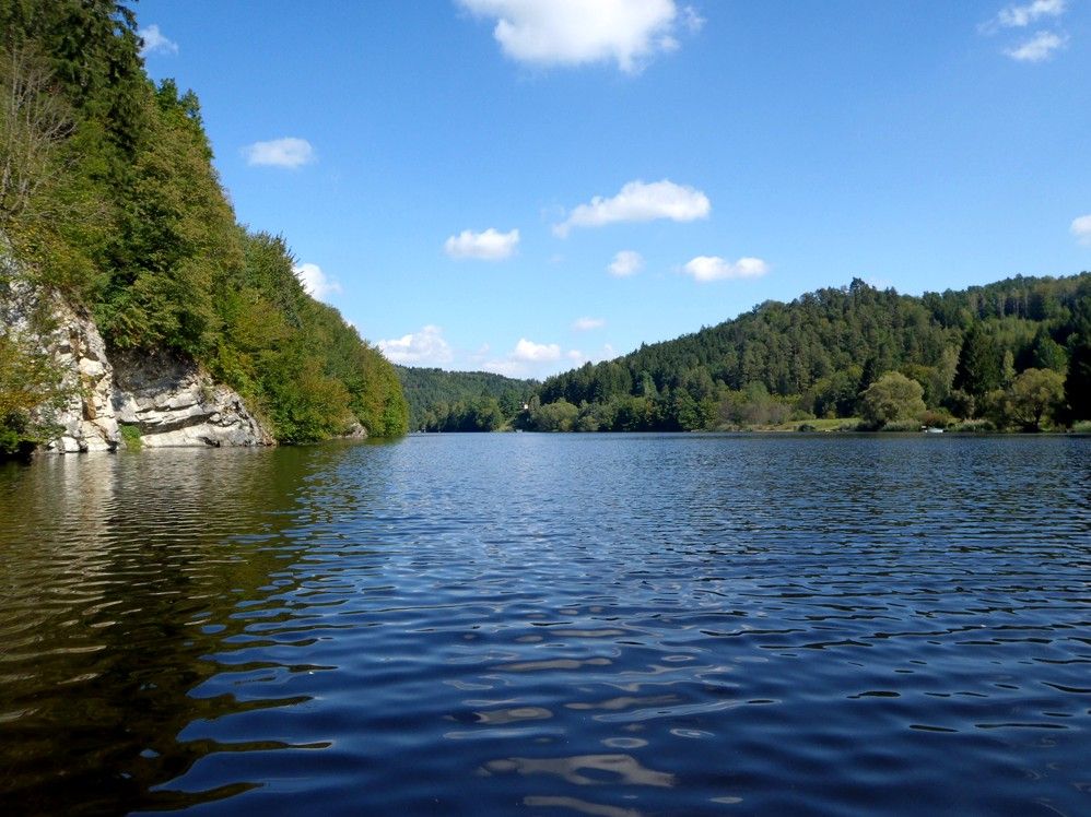 A calm lake surrounded by wooded hills under a blue sky with few clouds.