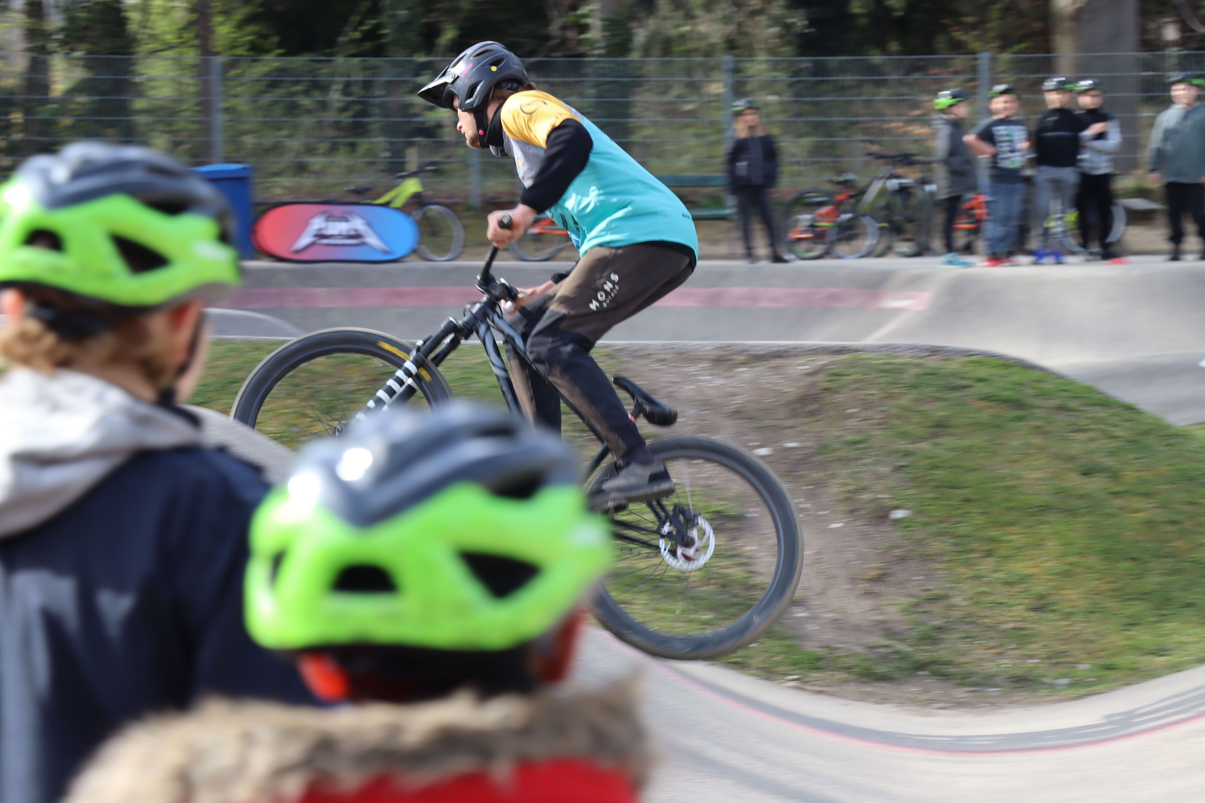 A cyclist jumps on a pump track while spectators with green helmets look on.