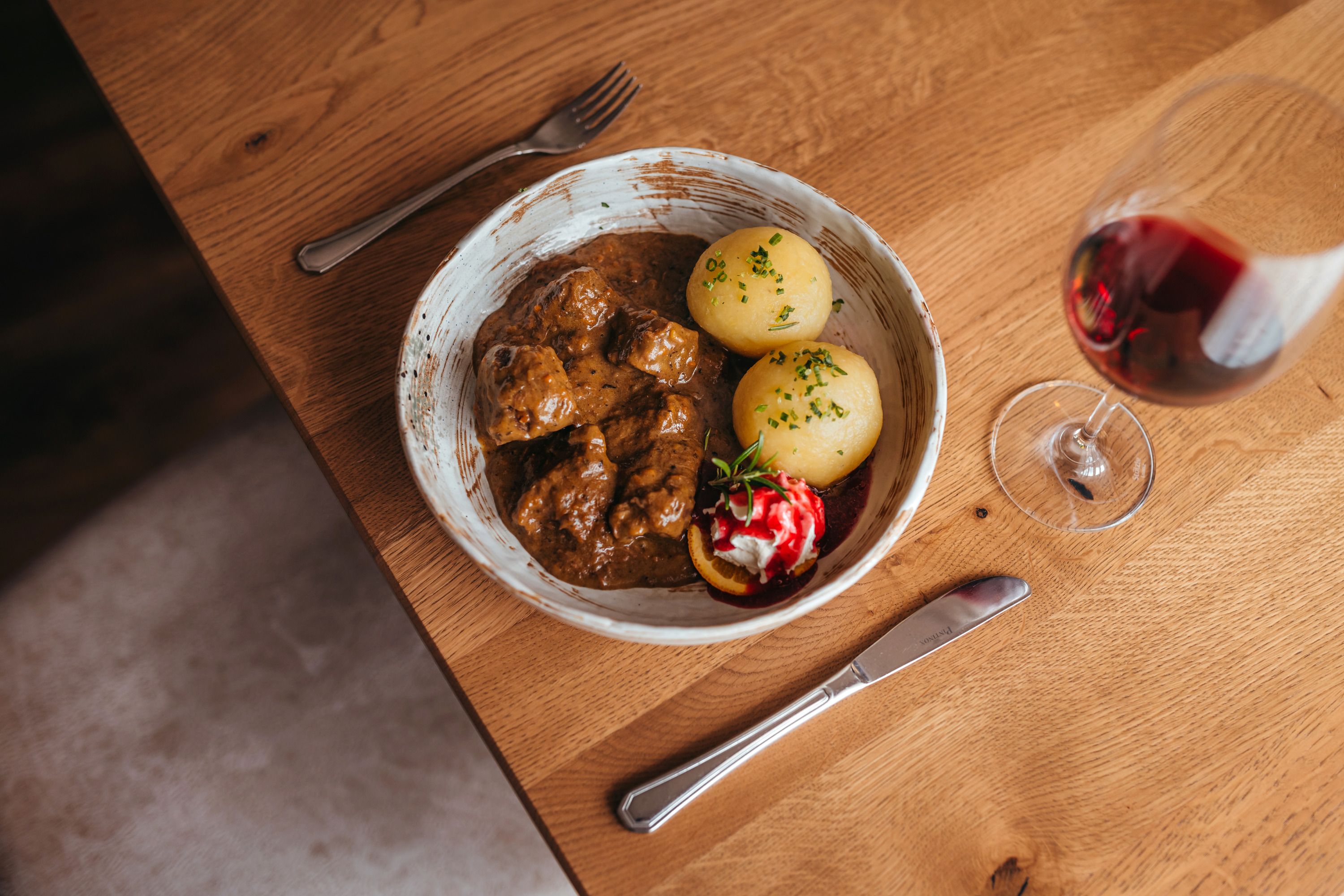 A plate of venison goulash, dumplings and cranberries, next to a glass of red wine on a wooden table.