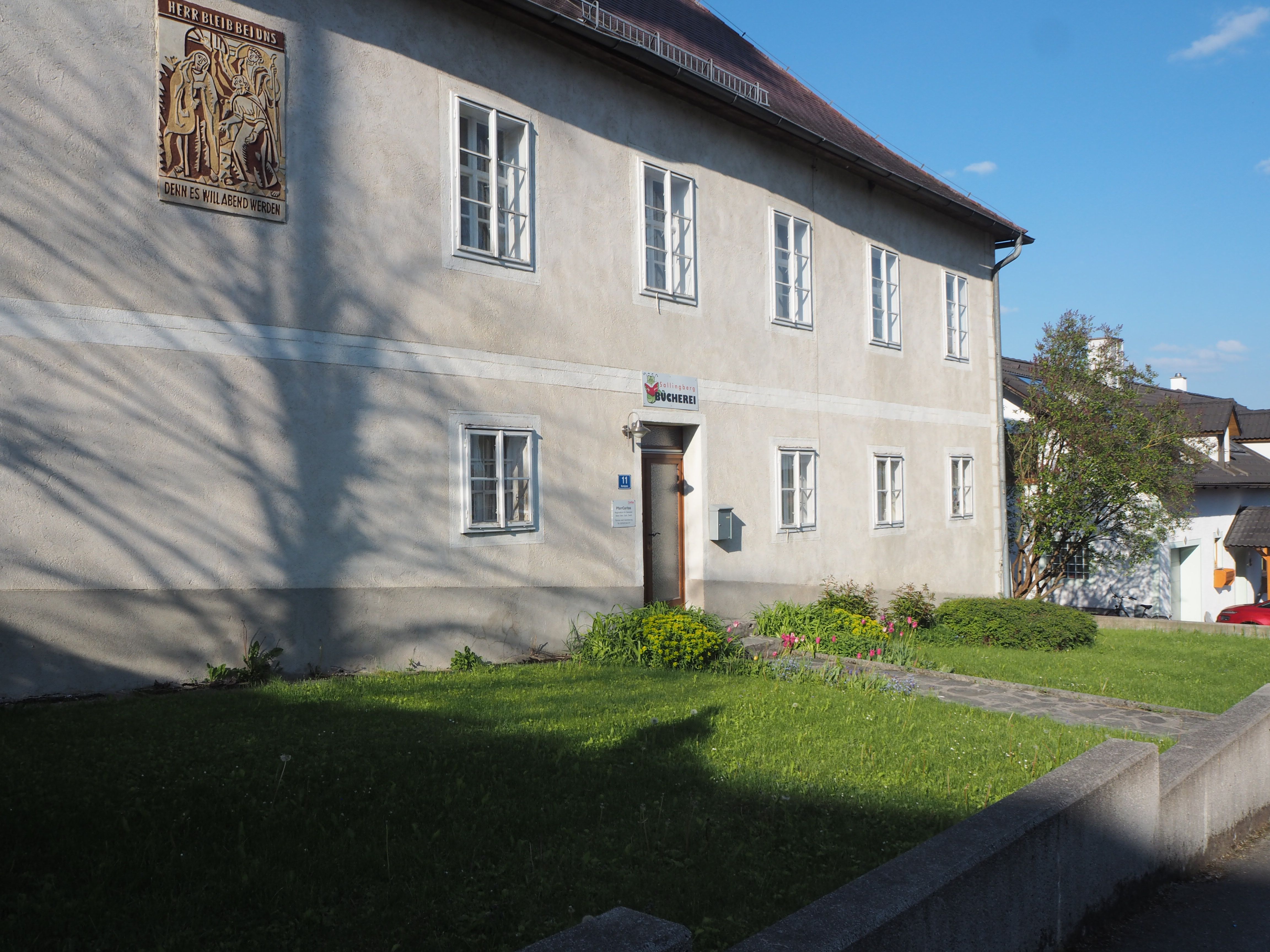 A two-story building with several windows and a sign above the door, surrounded by a well-tended garden.