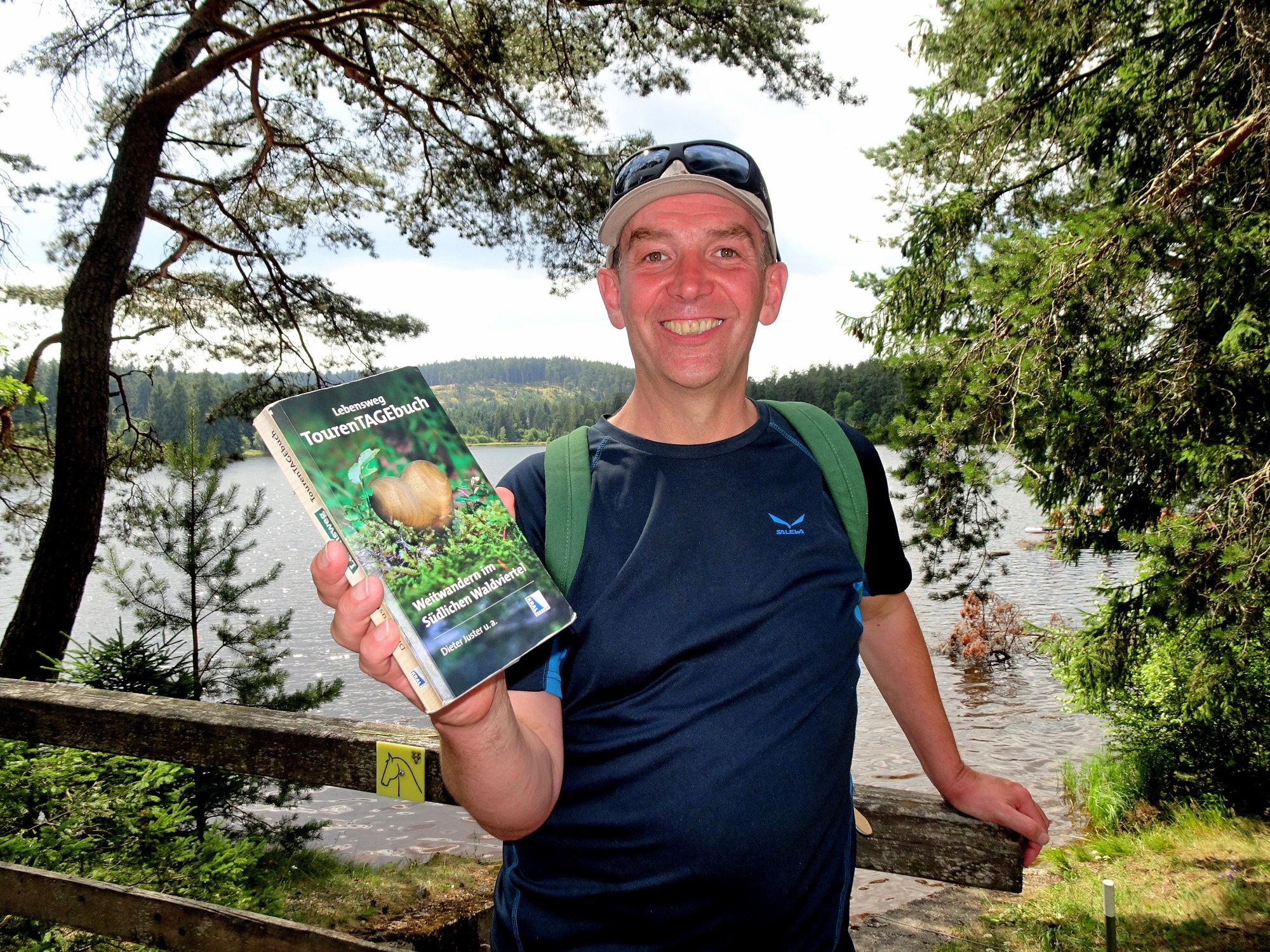 A man stands smiling in front of a lake, holding a book entitled 'Lebensweg Tourentagebuch'.