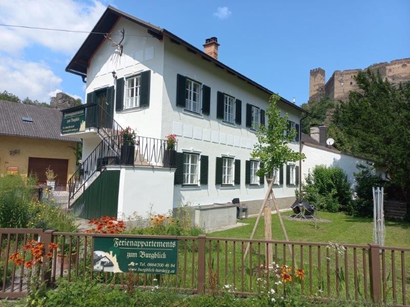 A white building with green shutters, surrounded by a garden, with a castle in the background.