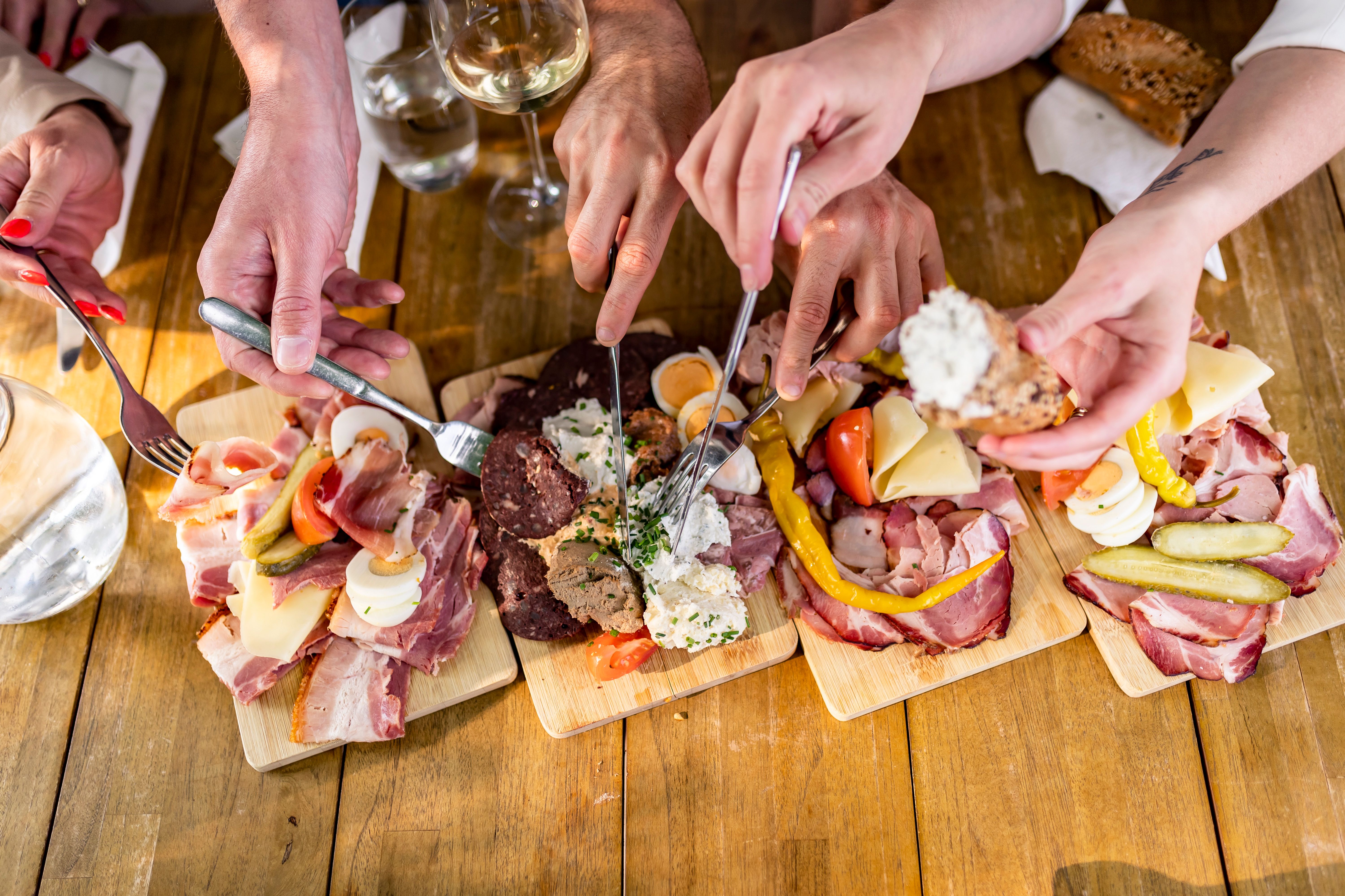 Several hands reach for platters of sausage and cheese on a wooden table.