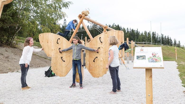 Children play on a large wooden butterfly in an outdoor playground.