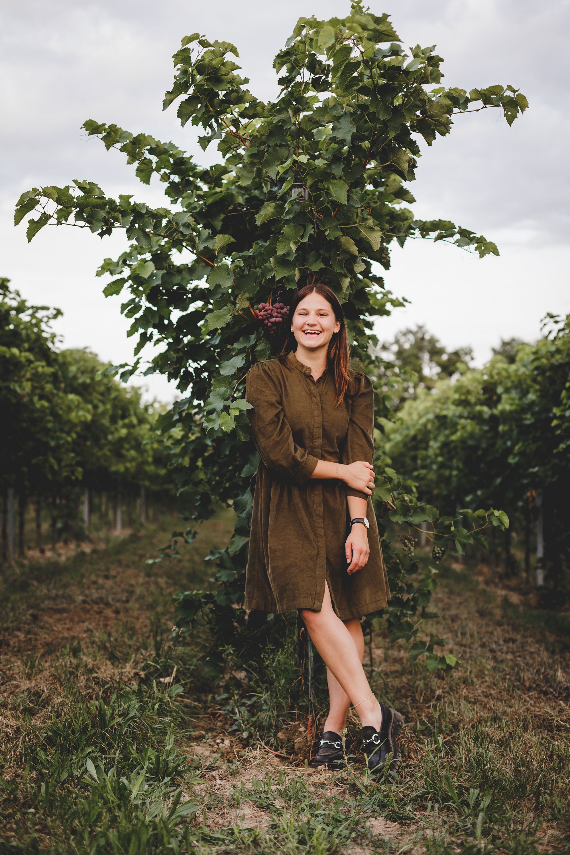 A woman stands smiling in a vineyard in front of a grapevine.