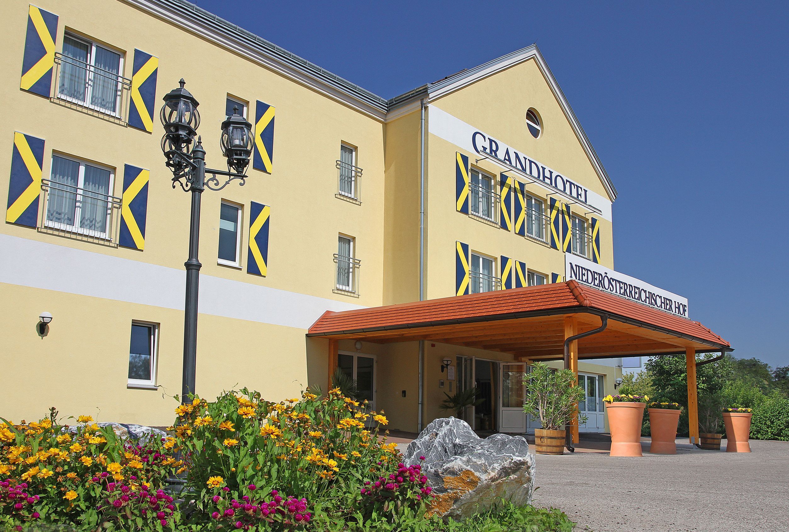 Exterior view of a yellow hotel with blue shutters and a canopy. Yellow and pink flowers bloom in front of the building.