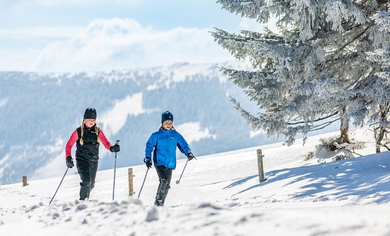 Two people cross-country skiing on a snow-covered trail with mountains in the background.