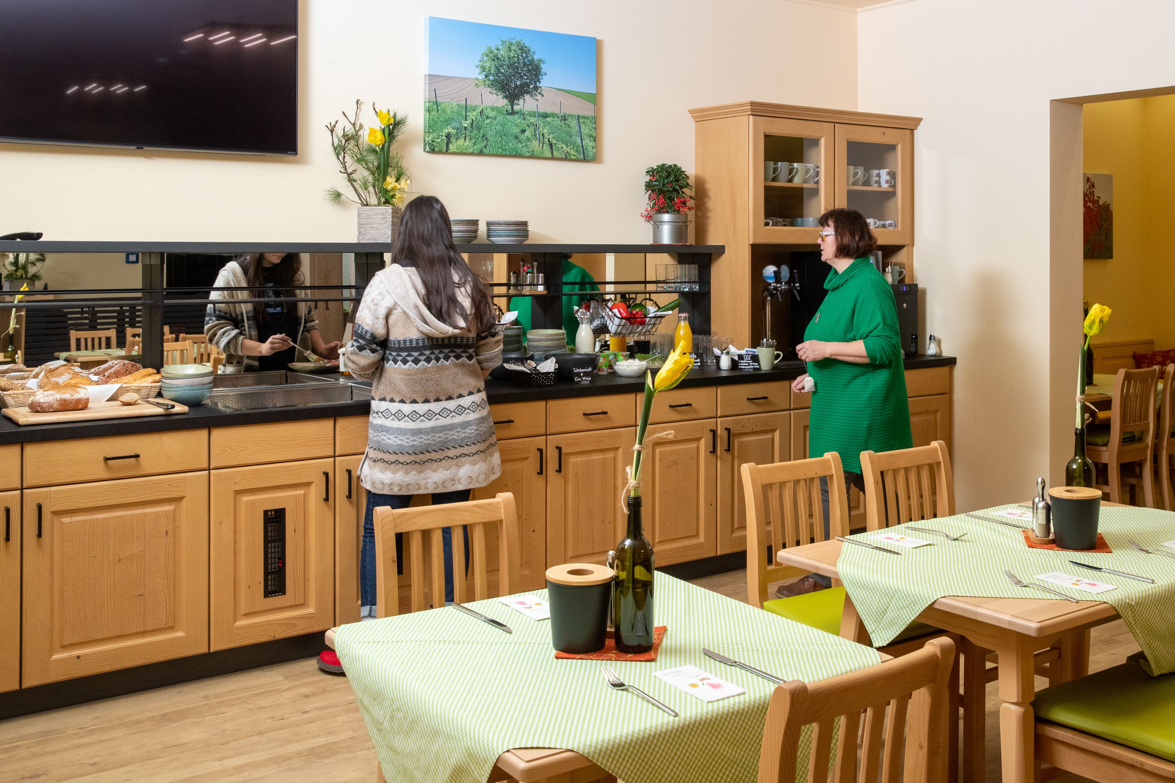 Two people help themselves to a breakfast buffet in a cozy room with wooden furniture and green tablecloths.