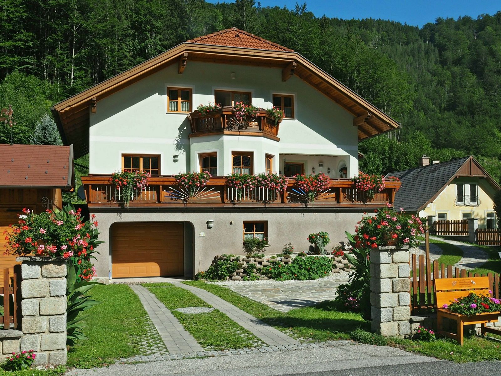 A two-storey house with wooden balconies and flowers, surrounded by green countryside.