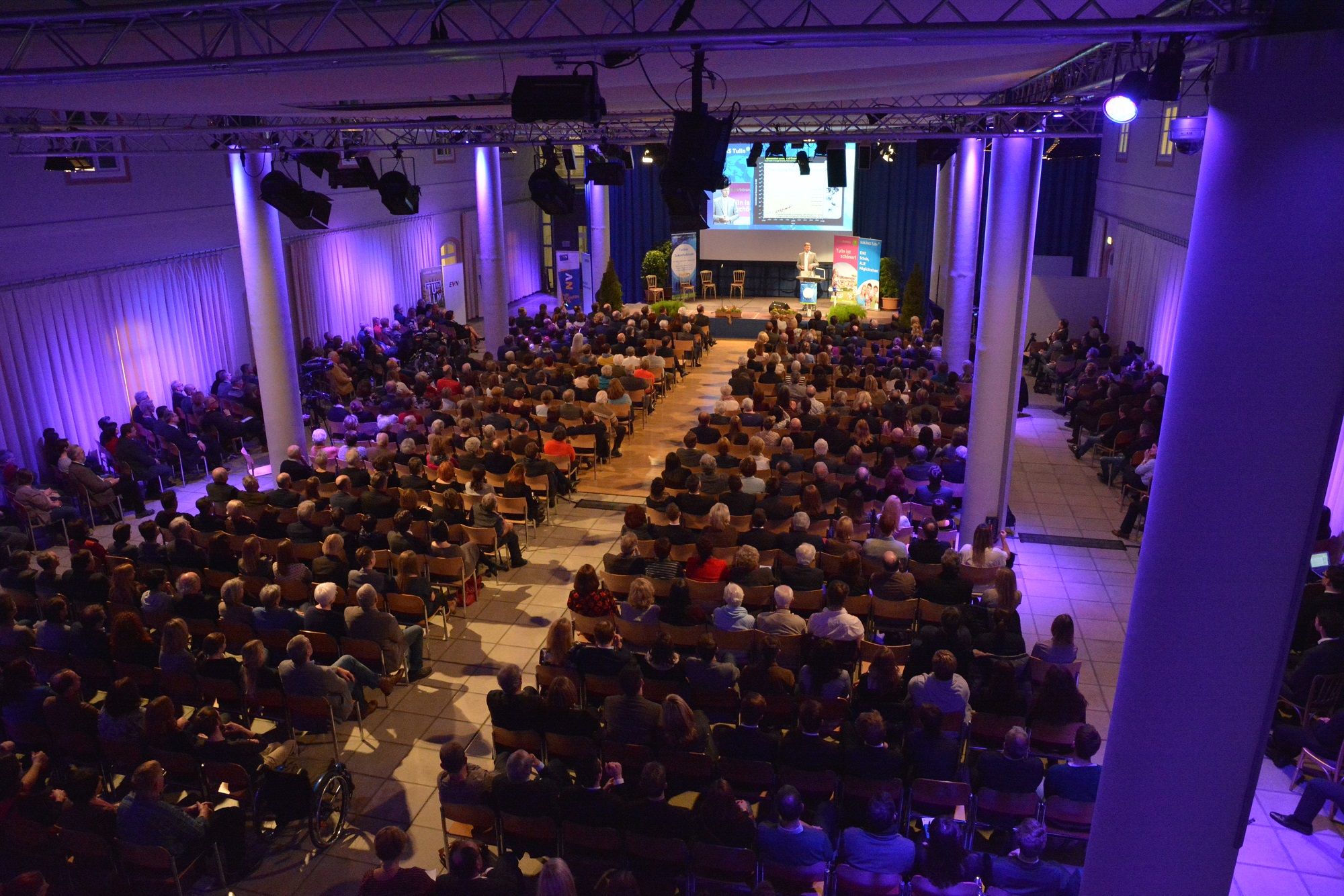 A fully occupied event room with stage and audience, illuminated in blue light.