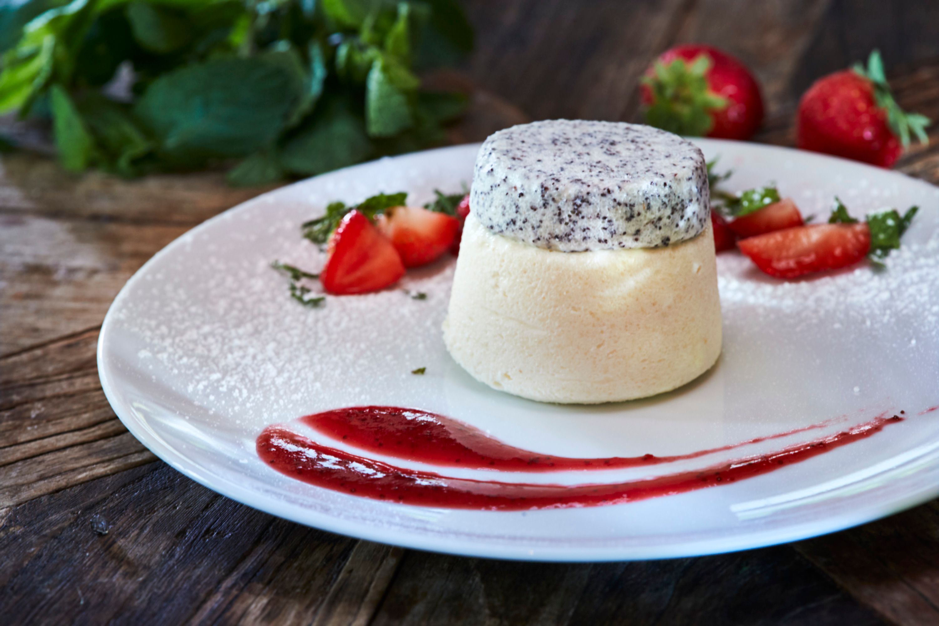 A dessert of semolina flummery topped with poppy seeds, garnished with strawberries and mint on a white plate.