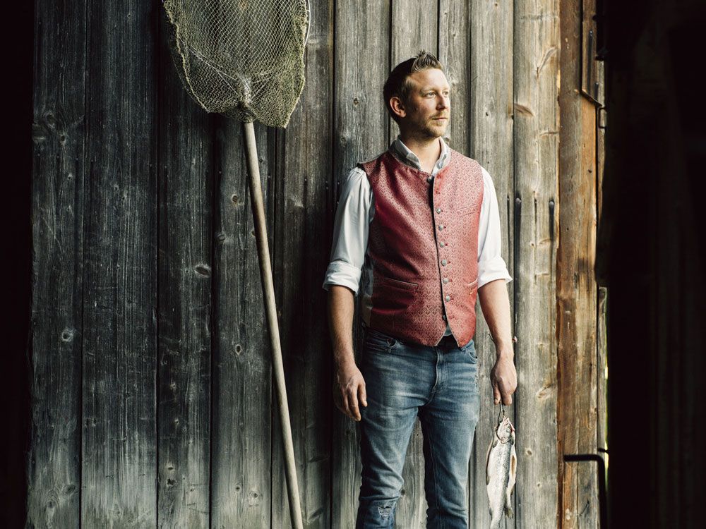 Man in red vest standing in front of wooden wall with fishing net and fish.