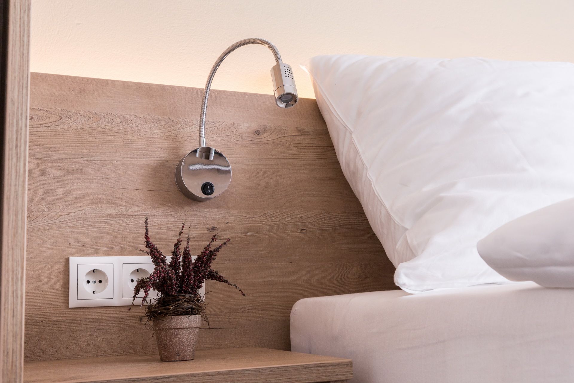 Close-up of a bed with wooden back panel, reading lamp, sockets and potted plant.