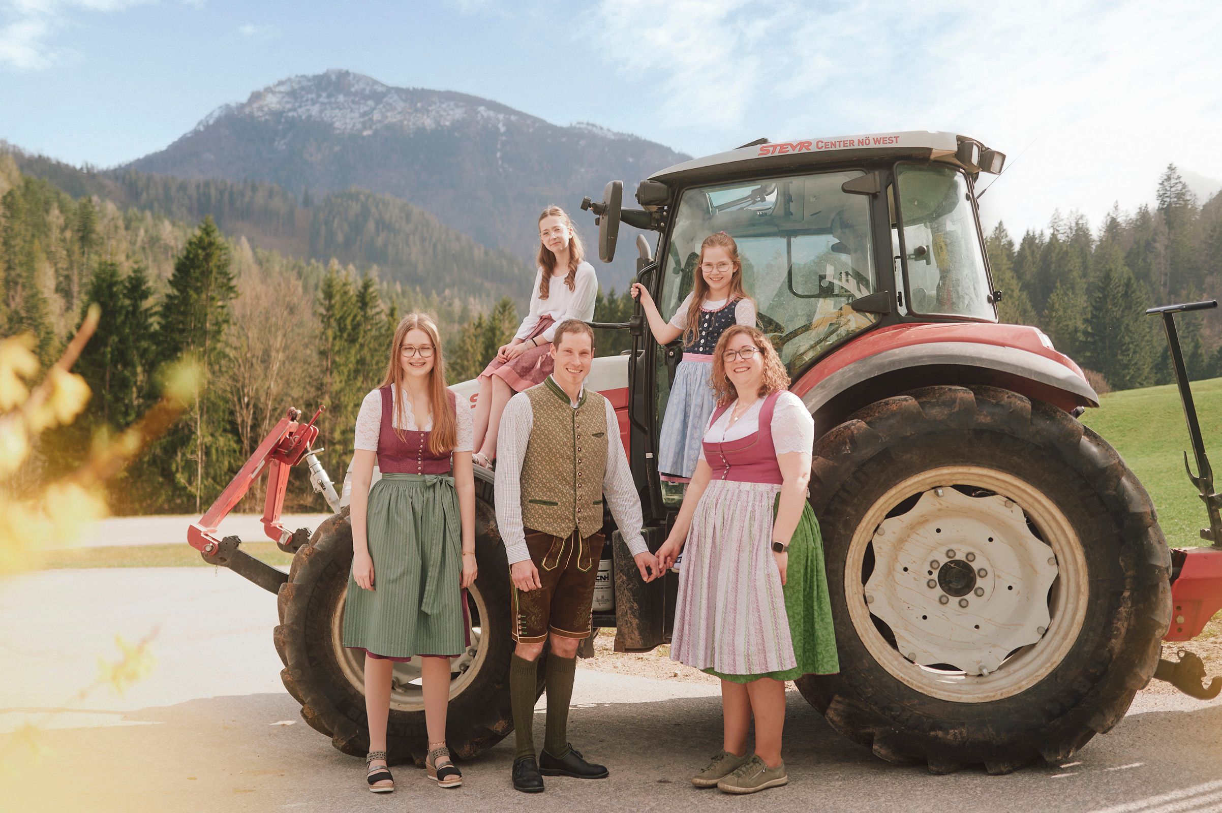 A family in traditional costume stands in front of a tractor in a rural mountain landscape.