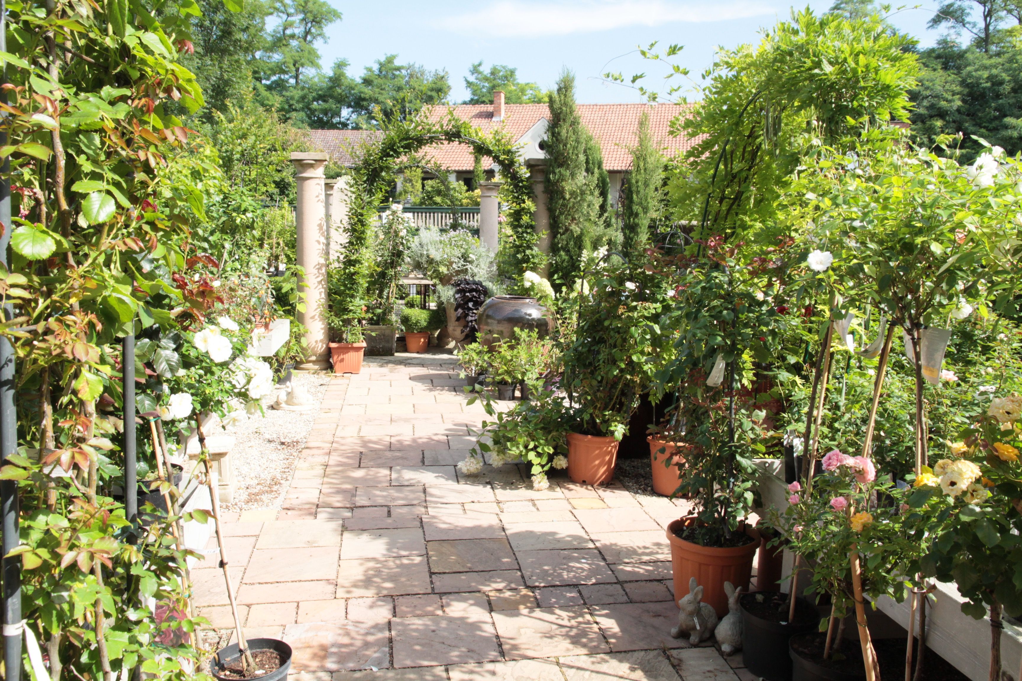 A well-kept garden path with potted plants and flowers, flanked by columns and an archway, leads to a building with a tiled roof.