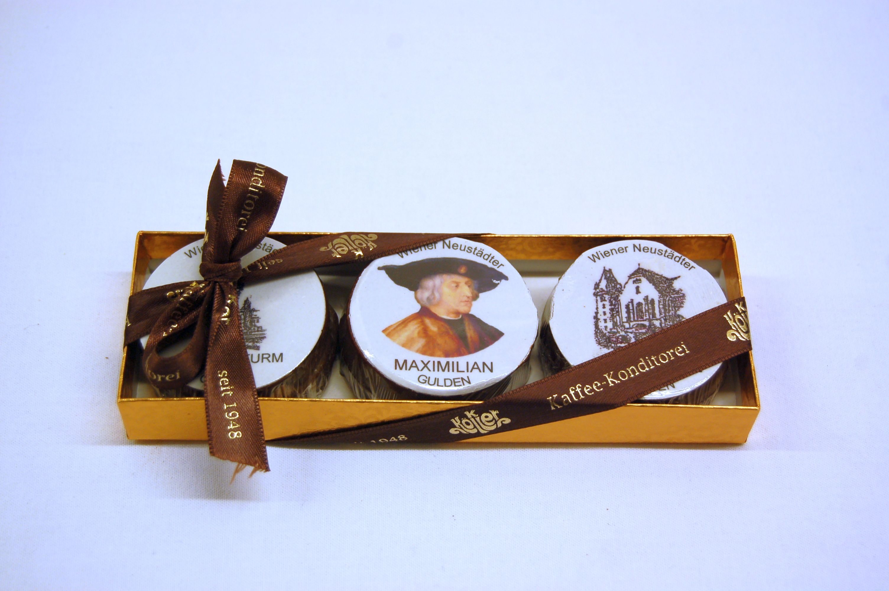 A golden box with three chocolates, decorated with a picture of Maximilian and Wiener Neustadt motifs, tied with a brown ribbon.