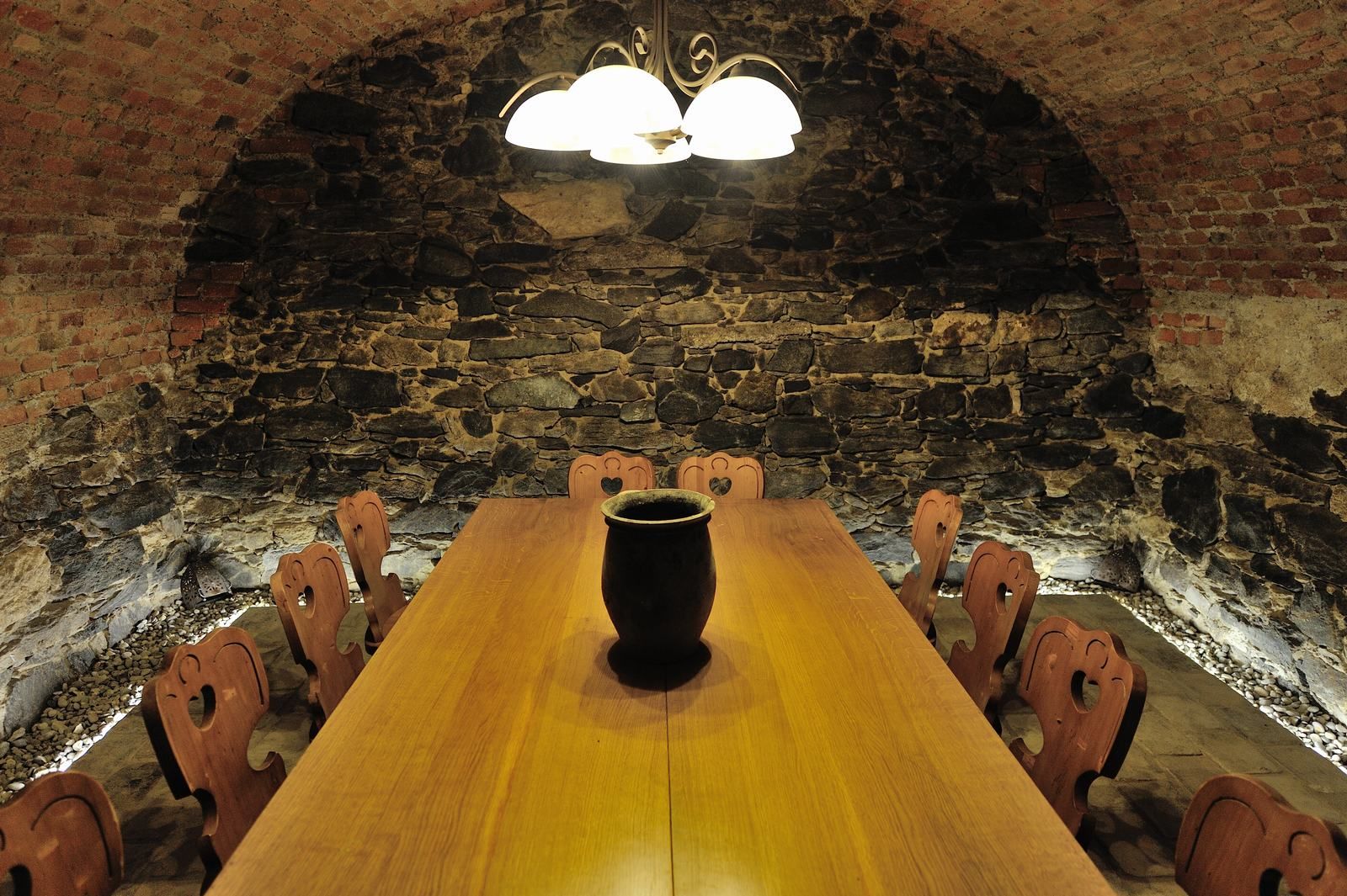 A rustic wine cellar with a long wooden table and chairs, surrounded by stone walls and illuminated by a ceiling lamp.