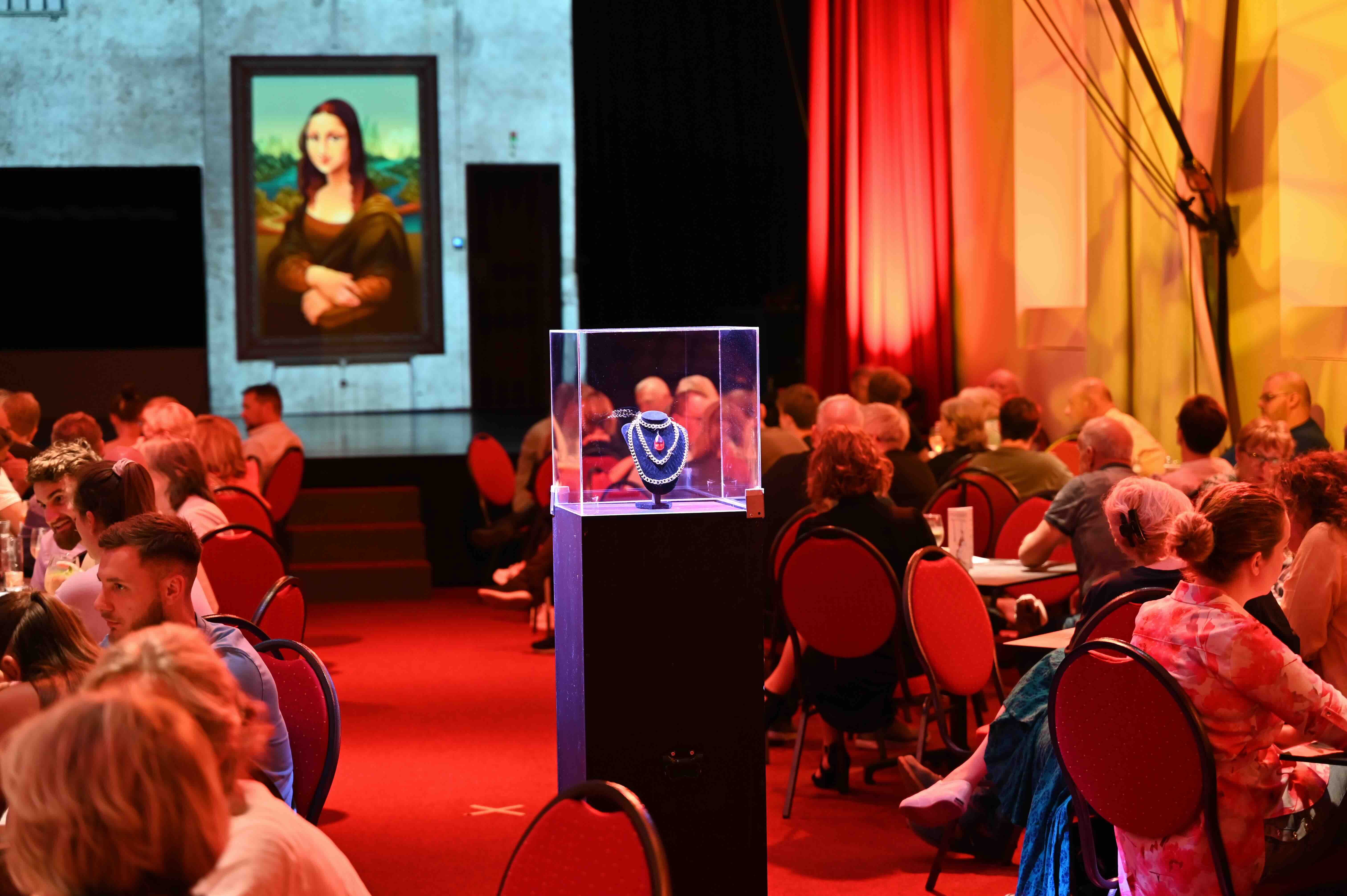 A theater with people at tables, an illuminated piece of jewelry in a display case and a picture of the Mona Lisa in the background.