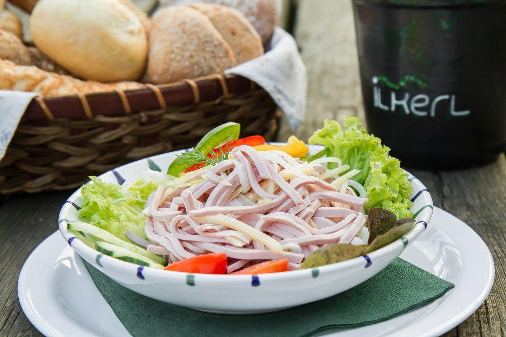 A plate of sausage salad, garnished with lettuce leaves, tomatoes and cucumber. A bread basket in the background.