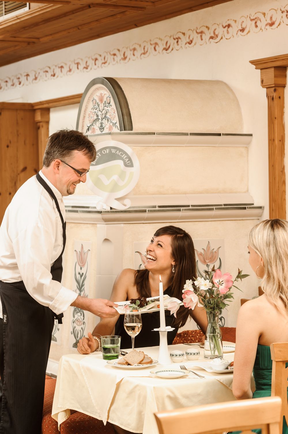 A waiter serves two women in a cozy restaurant. One woman laughs as she accepts the food. The table is elegantly set.